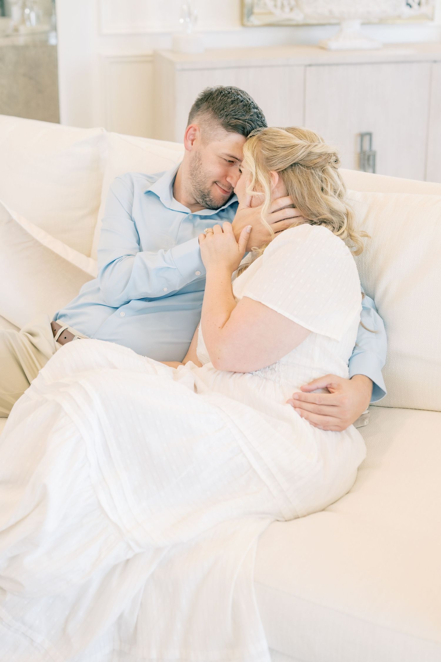 A couple in wedding attire shares a romantic moment on a white sofa, with the groom tenderly holding the bride's face.