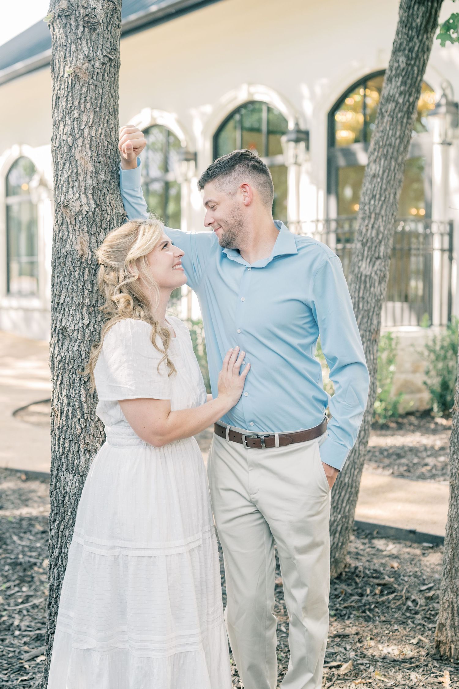 Laughing couple standing between two trees outside a white building with gothic arched windows on a sunny day.
