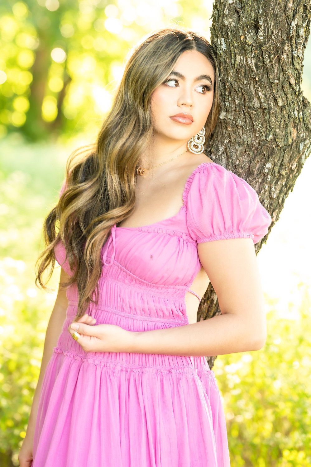 Young woman in pink ruched dress with puff sleeves poses by tree in sunlit garden.