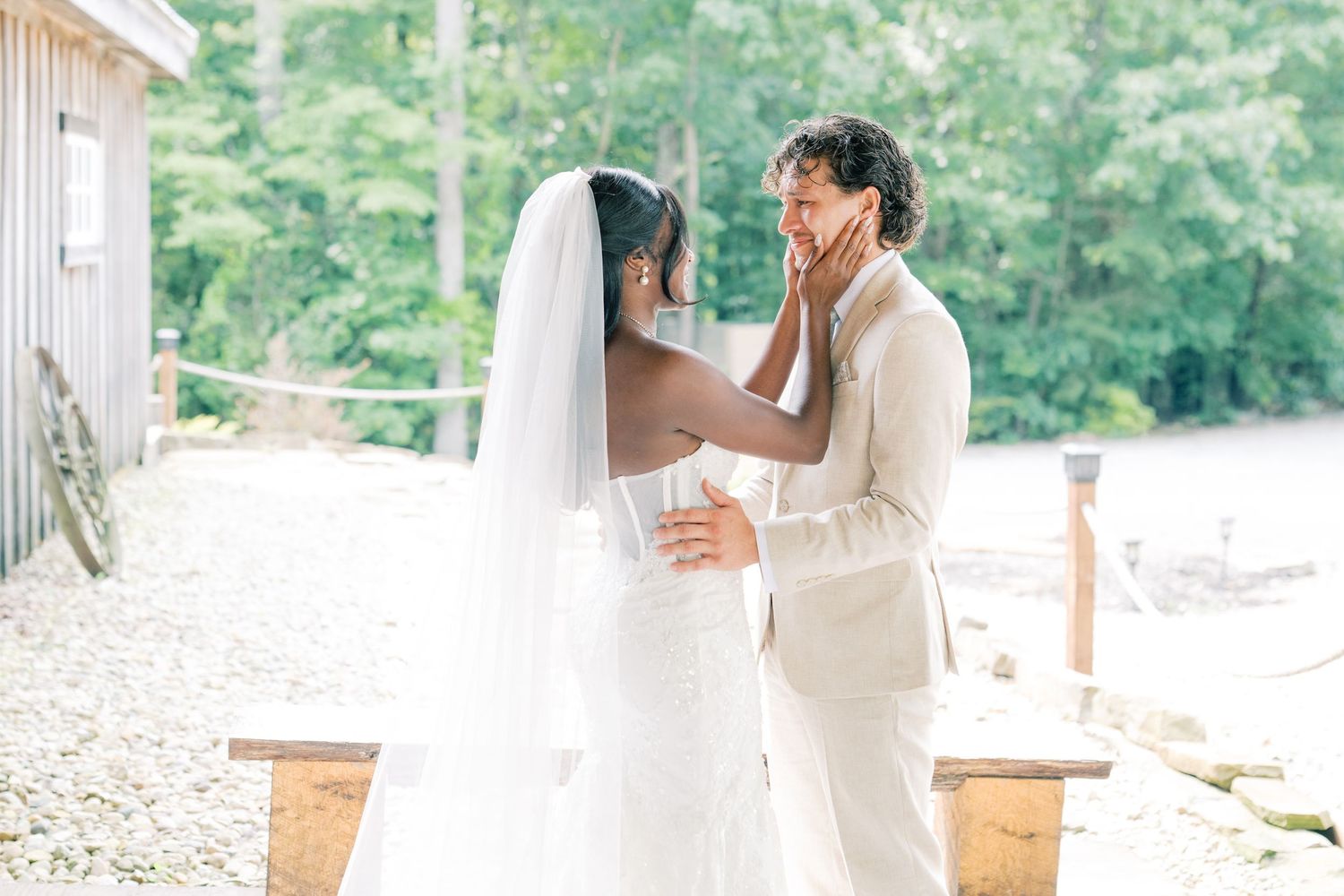 Bride and groom share an emotional first look moment outdoors, with bride in white gown and veil, groom in tan suit.