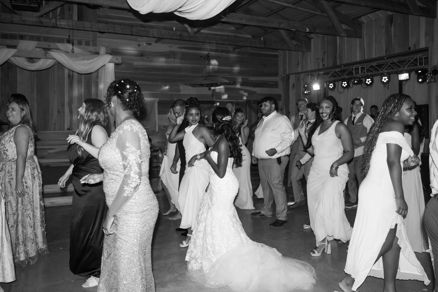 Bride in white gown dances joyfully with wedding guests on crowded reception dance floor in black and white photo.