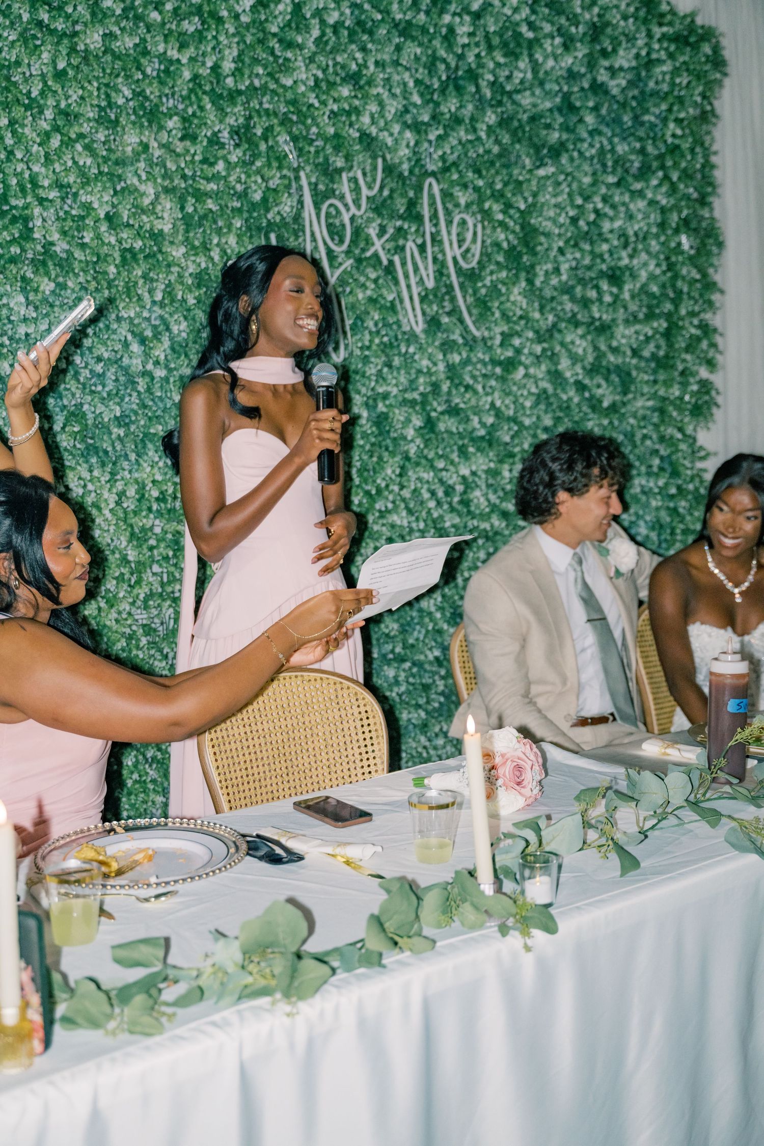 Bridesmaid gives toast at wedding reception in front of green floral wall with love sign.