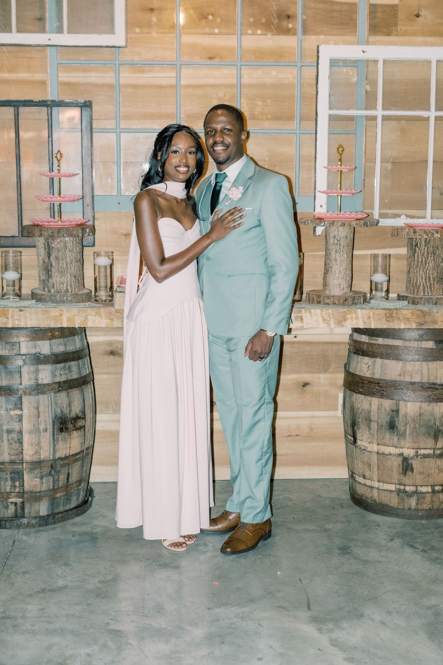 Couple poses together at wedding reception, woman in blush gown and man in mint suit near rustic wooden barrels.