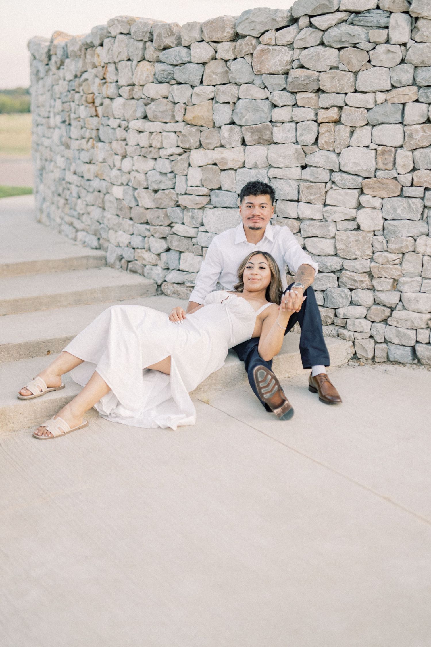 A laughing couple sits casually against a rustic stone wall during their relaxed outdoor engagement shoot.