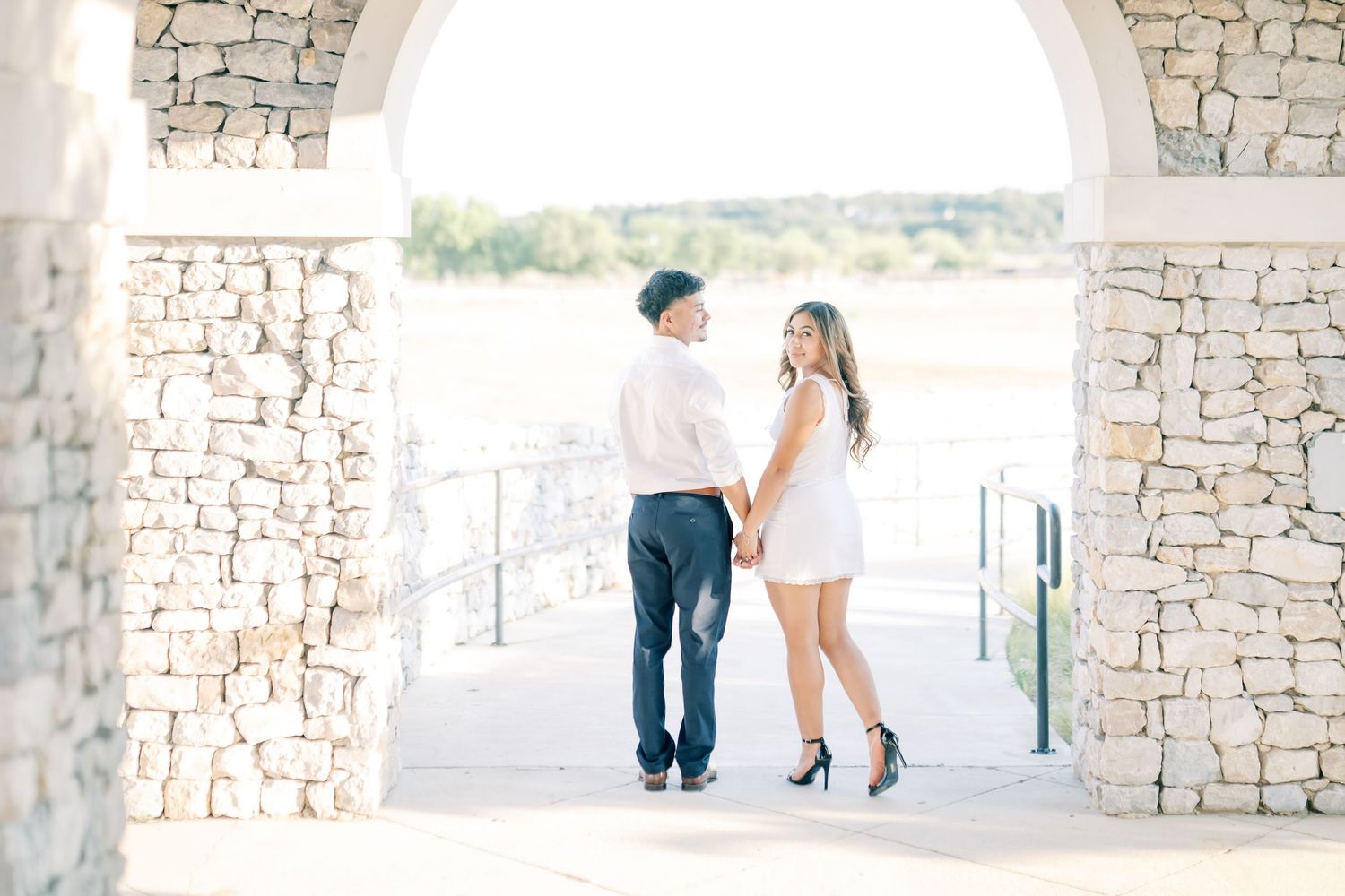 Couple holding hands under stone arch, woman in white dress glancing back, man in navy pants and white shirt, outdoor setting.
