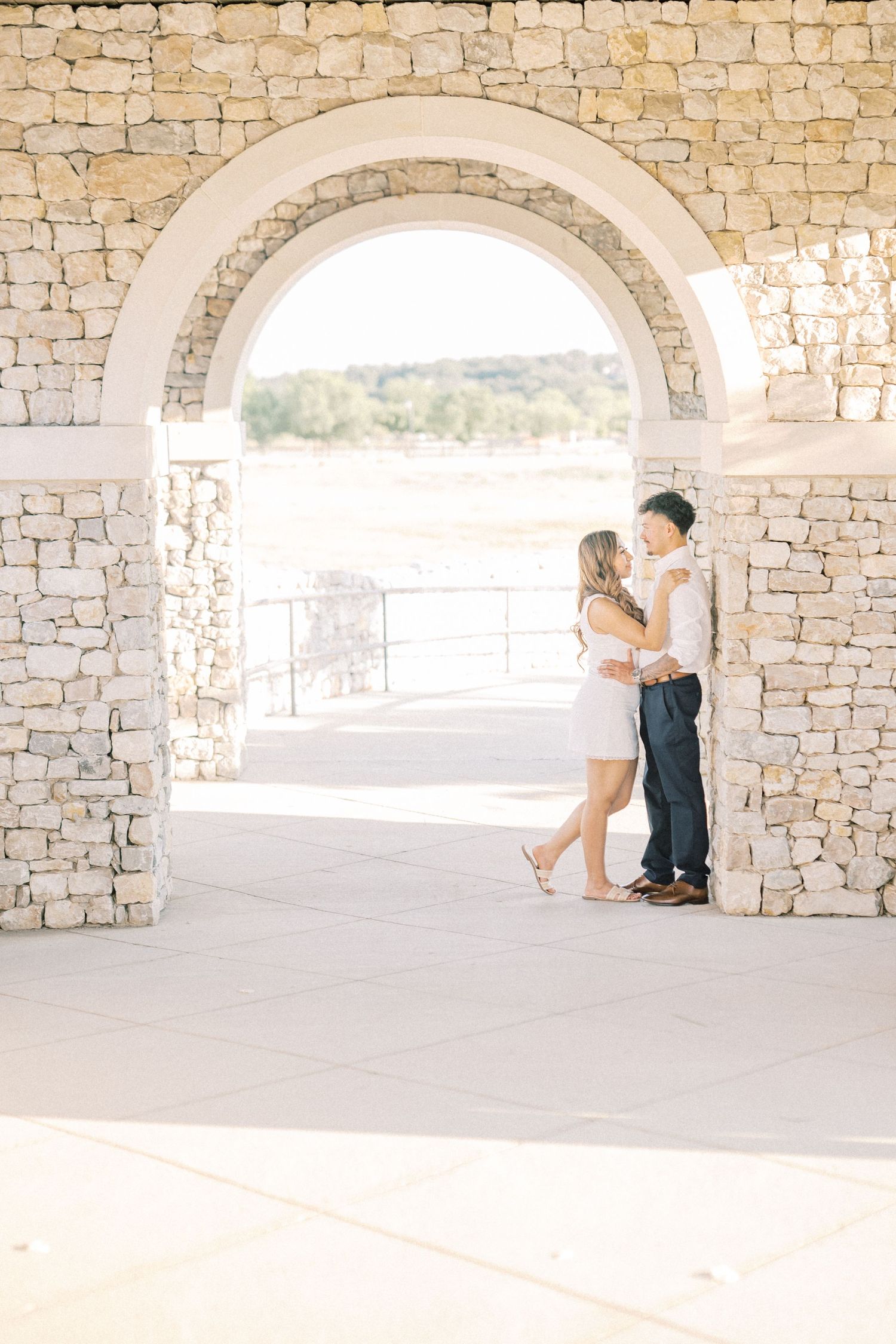 Couple embracing under a stone archway at an outdoor venue with scenic views in the background.
