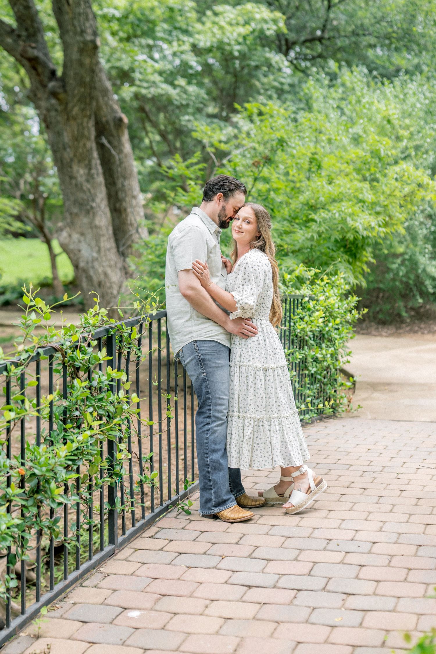 Couple embracing and smiling by iron fence in lush green park, woman in floral dress, man in casual shirt and jeans.