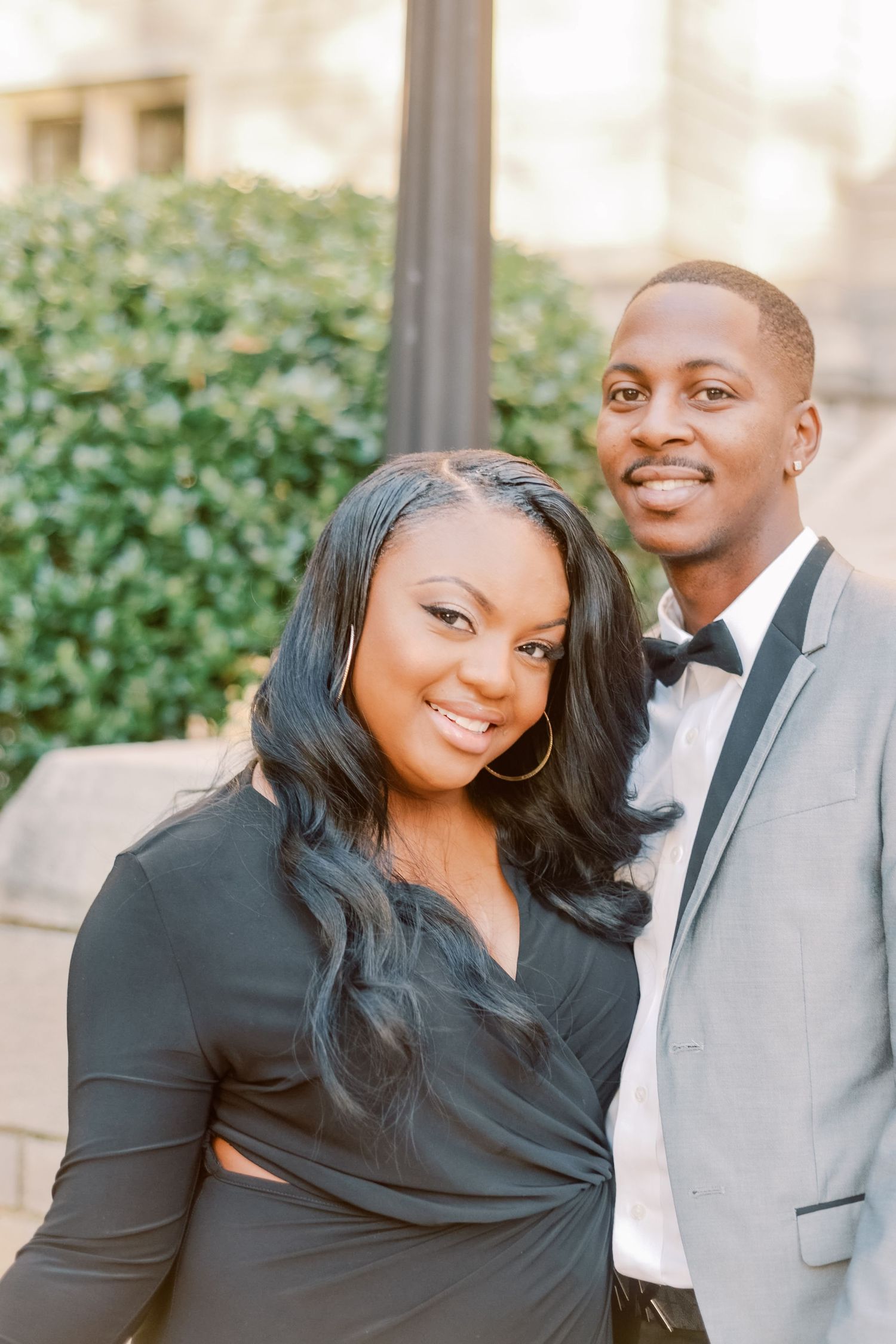 A smiling couple dressed elegantly in black outfits poses outdoors near green hedges for a formal portrait.