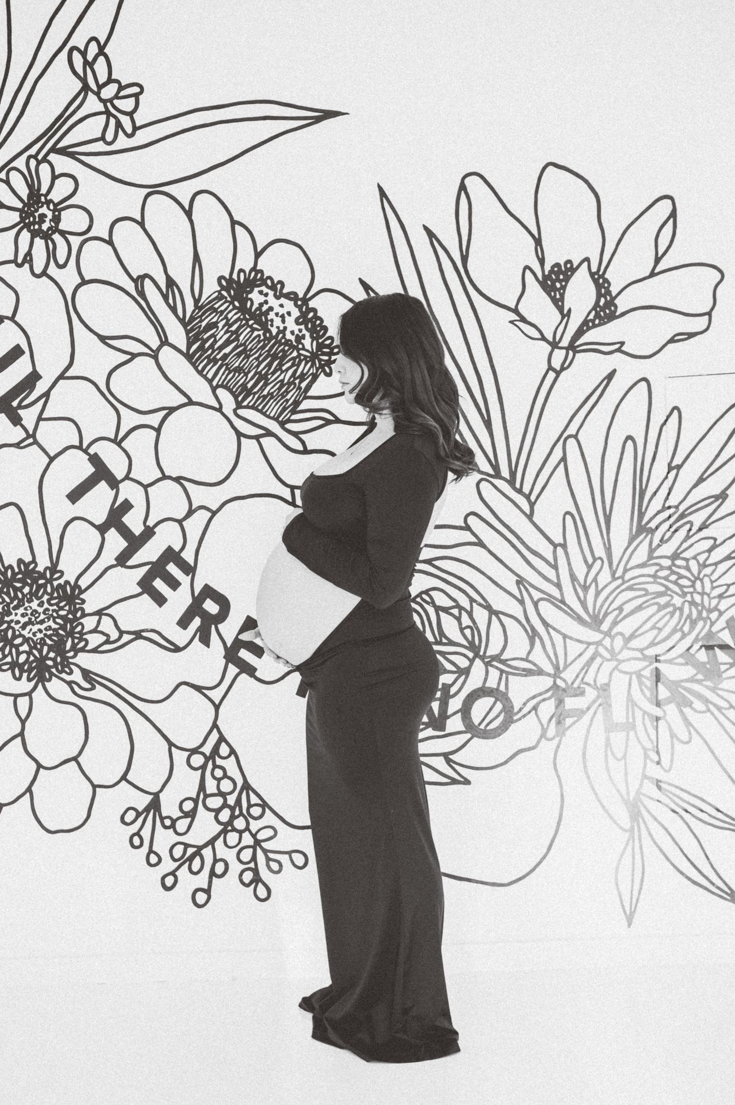 Pregnant woman in black dress posing sideways before floral mural wall art.