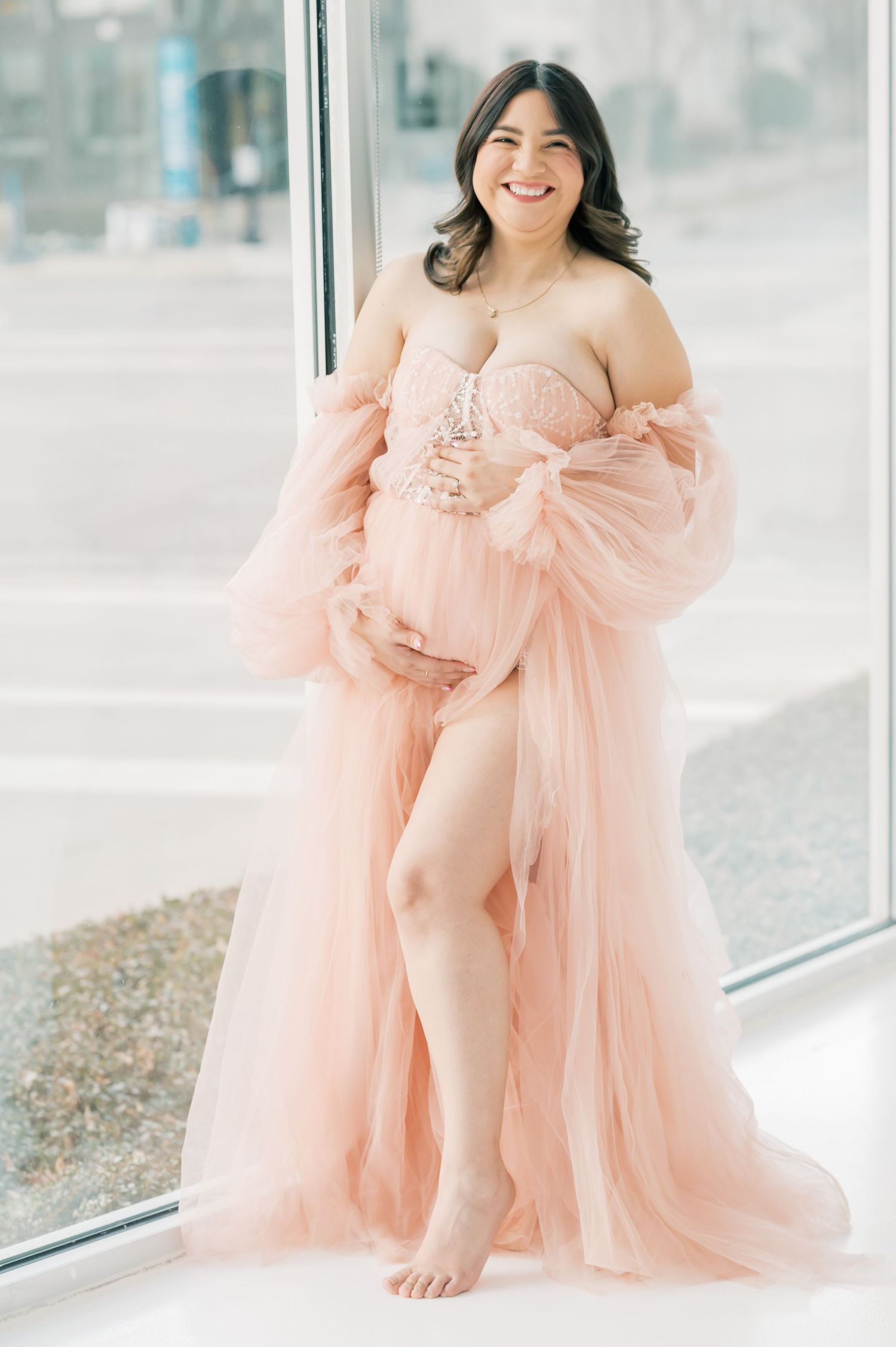 Smiling pregnant woman in a flowing blush pink tulle maternity gown posing by a large window in natural light.