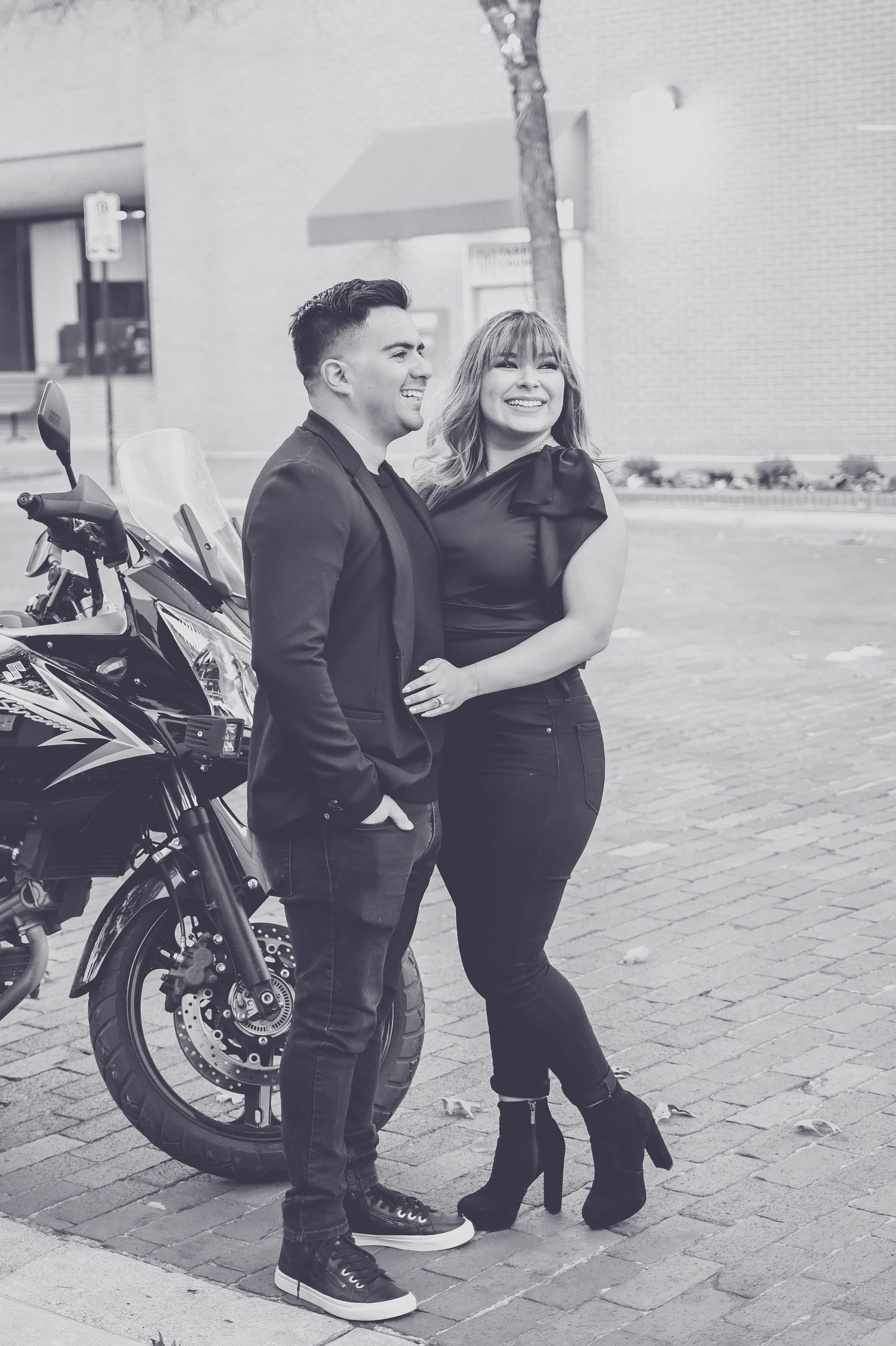 Couple posing in black outfits beside a motorcycle on a brick sidewalk in a black and white photo.