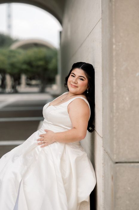 Bride in white ball gown leans against stone pillar outdoors, smiling gracefully during bridal portrait session.