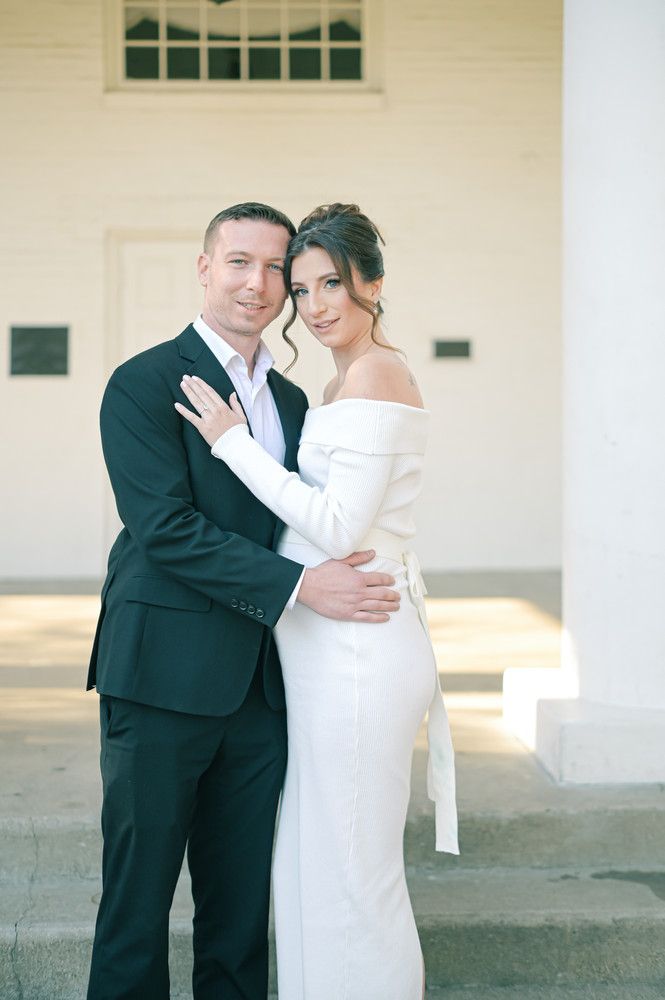 Elegant couple posing in wedding attire outside a white building, bride in off-shoulder gown.