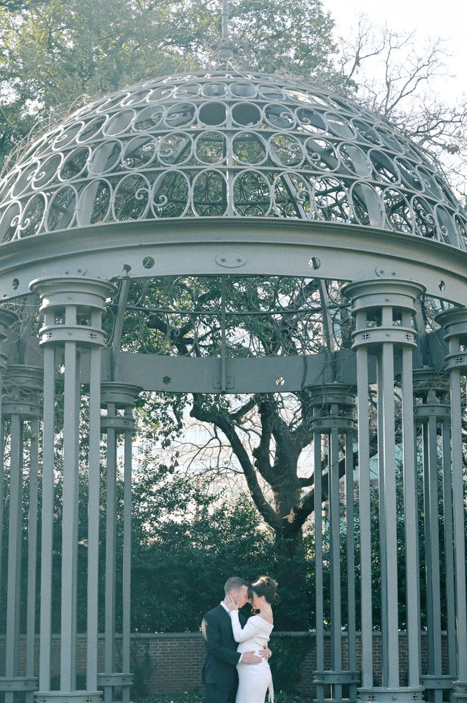 Romantic couple embracing under an ornate iron gazebo surrounded by trees during engagement photos.