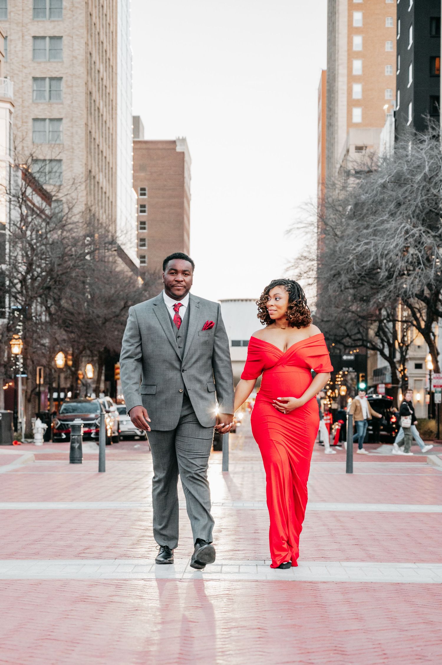 Pregnant couple walking hand-in-hand downtown, woman in red maternity gown and man in gray suit.