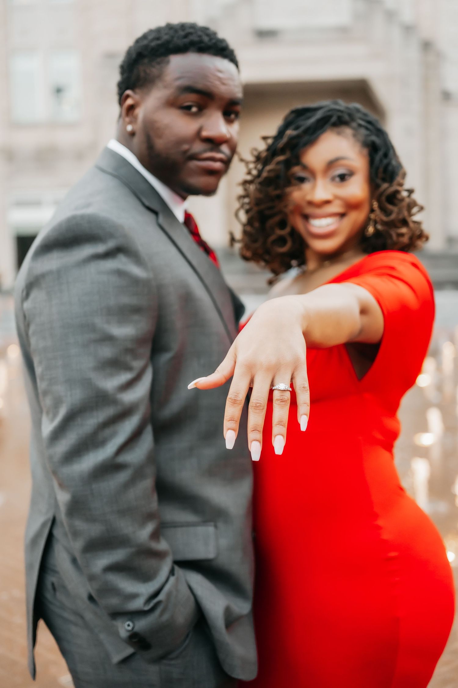 Smiling couple in red dress and gray suit showing off engagement ring outdoors.
