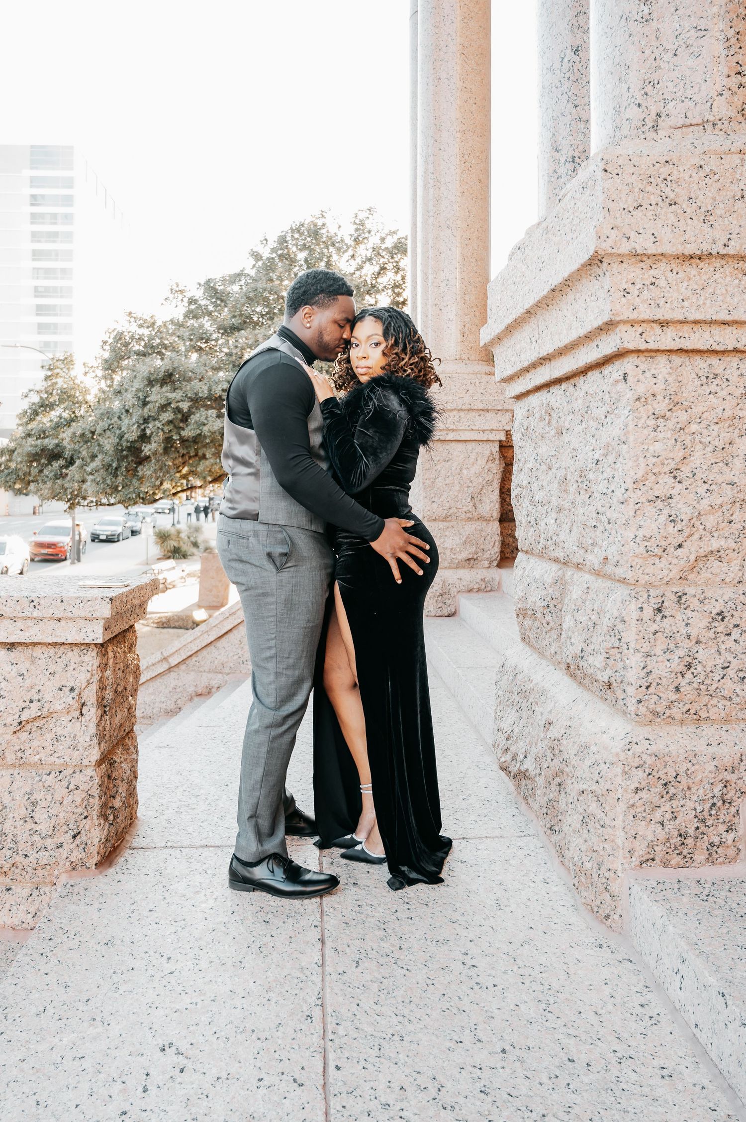 Elegant couple embracing near stone columns, man in gray suit and woman in black velvet gown, outdoor engagement photo.
