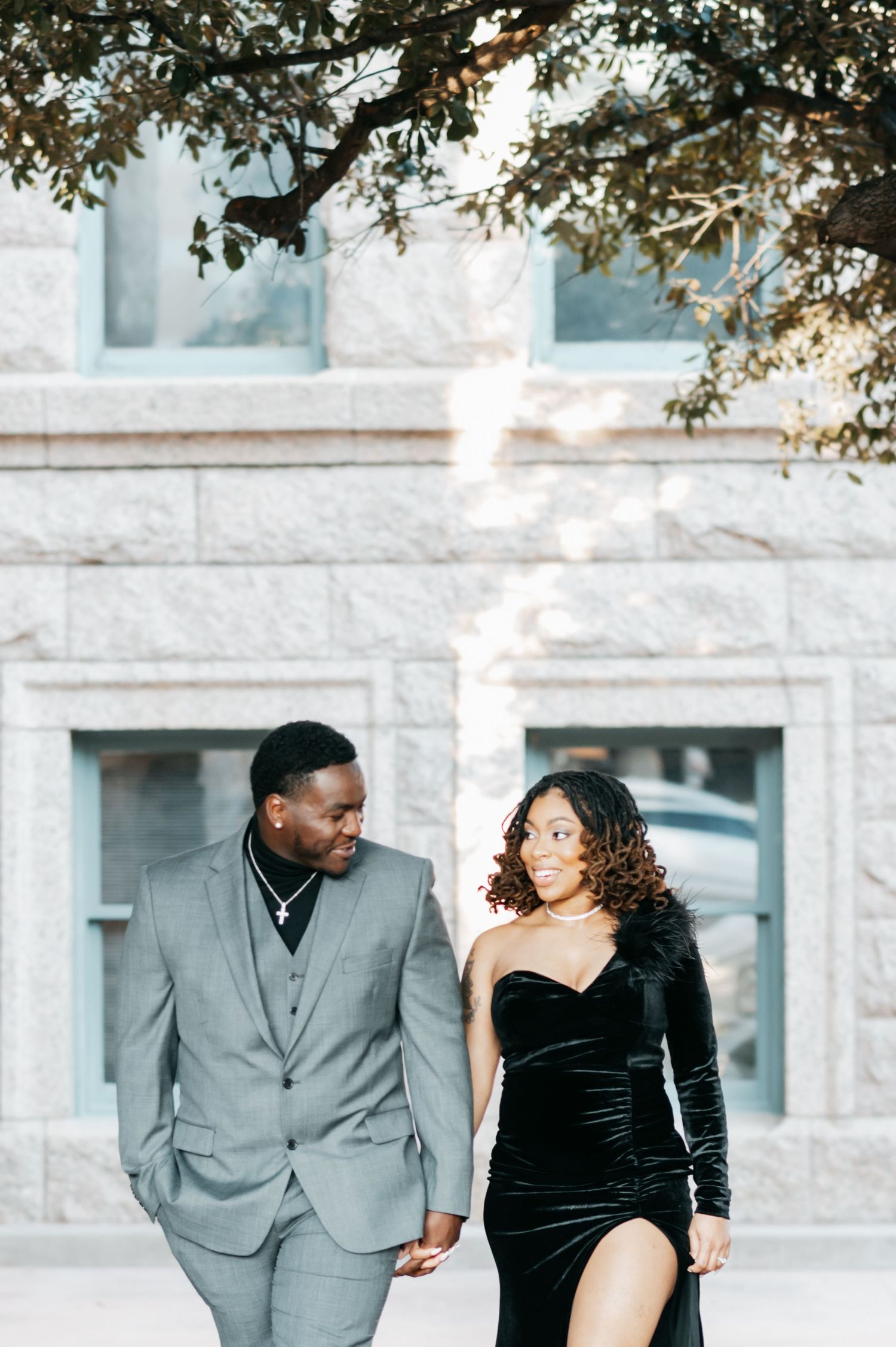 Elegant couple walking outdoors, man in gray suit and woman in black velvet gown, in front of a stone building.