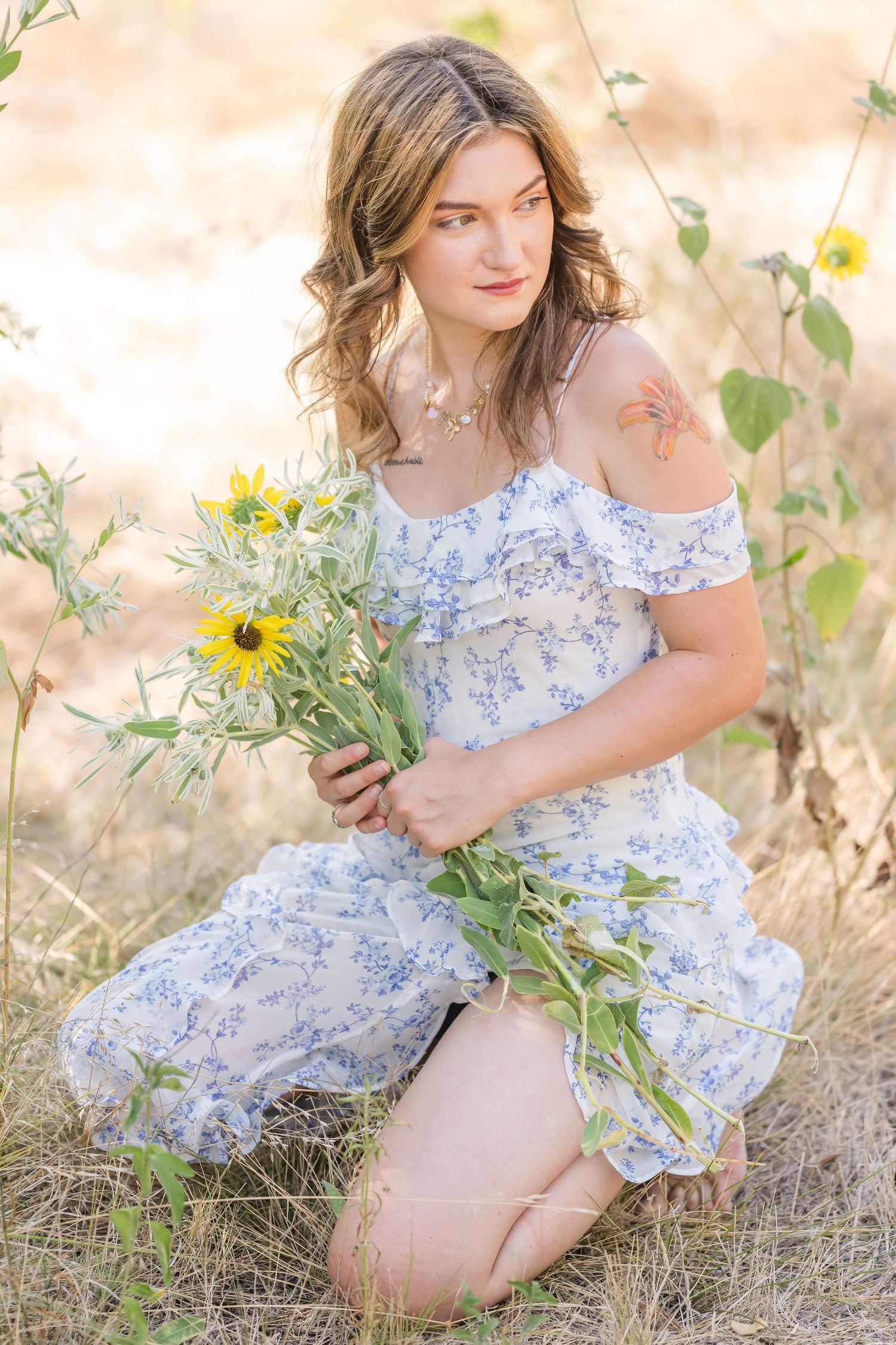 Young woman in blue floral off-shoulder dress kneeling in field holding sunflowers.