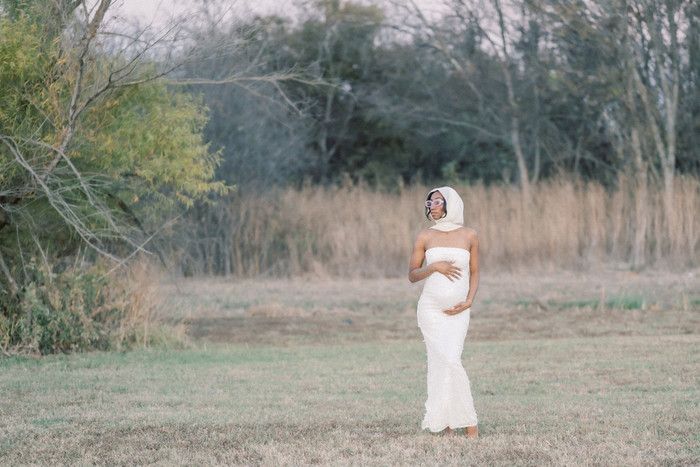 Wide landscape maternity photo showing woman in white dress standing in open field.