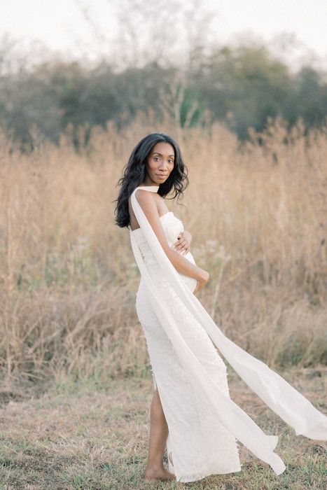 Artistic maternity portrait in white dress against natural field backdrop with flowing fabric.