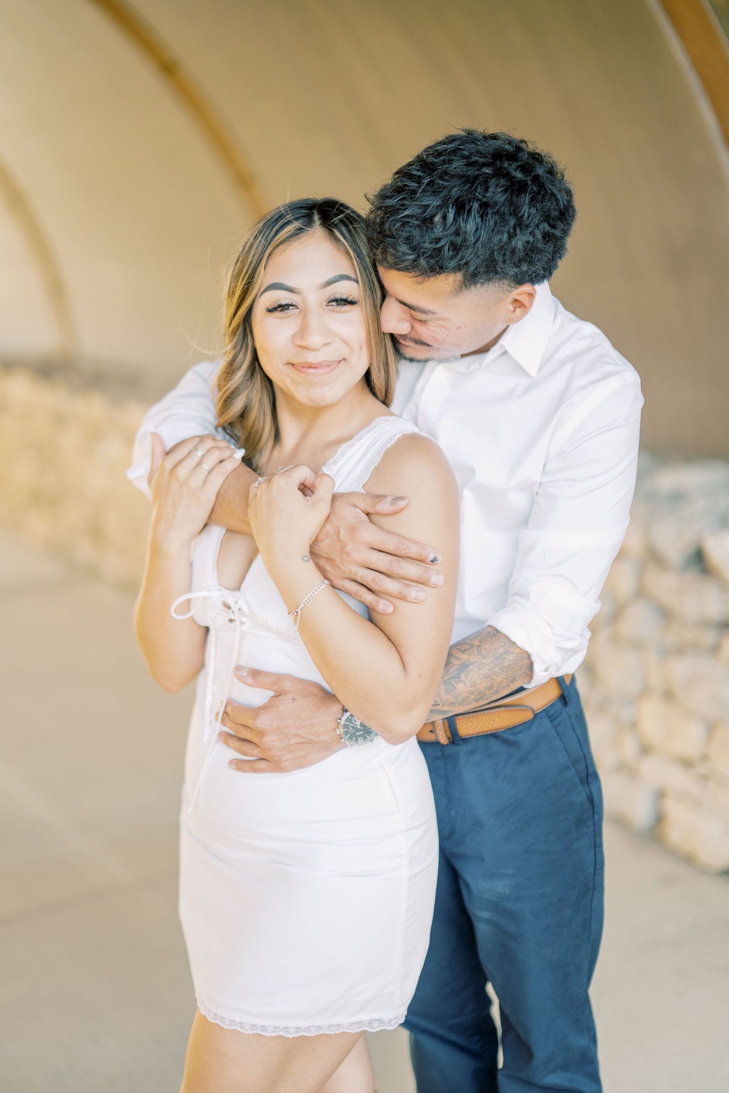 Couple embracing under an arch, woman in white dress smiling at camera while man holds her close.