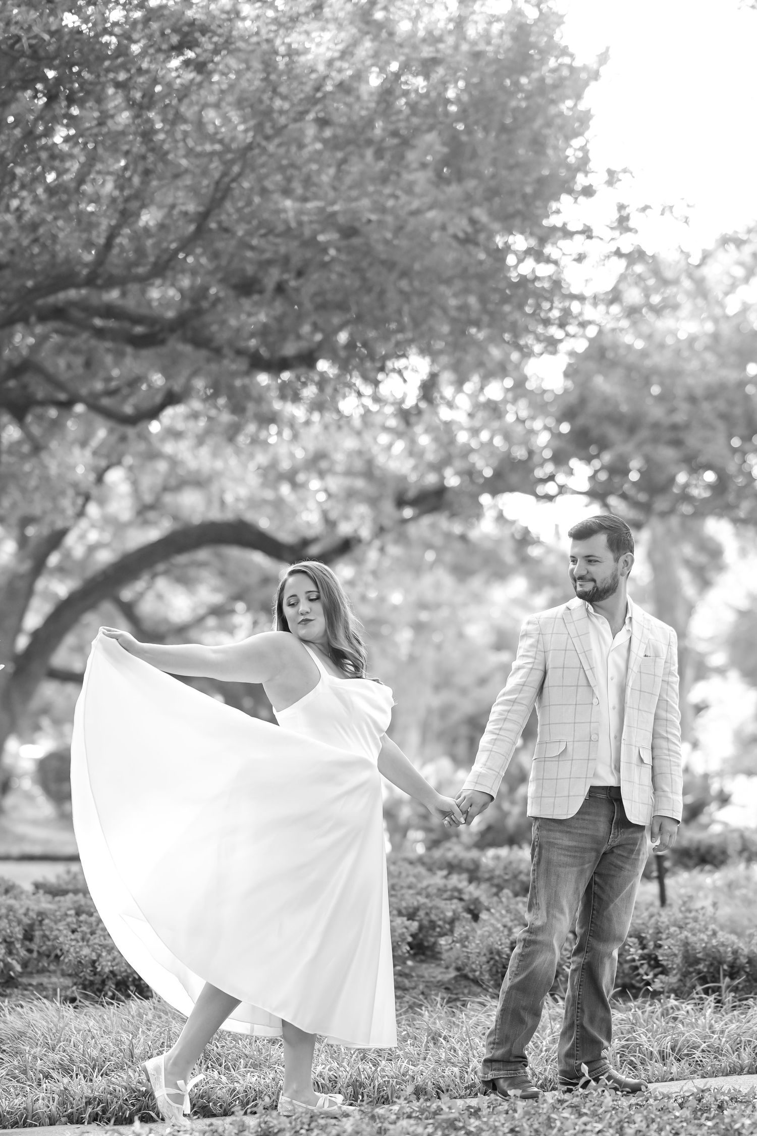Black and white photo of a woman in a flowing white dress twirling while holding hands with a smiling man under large park trees.