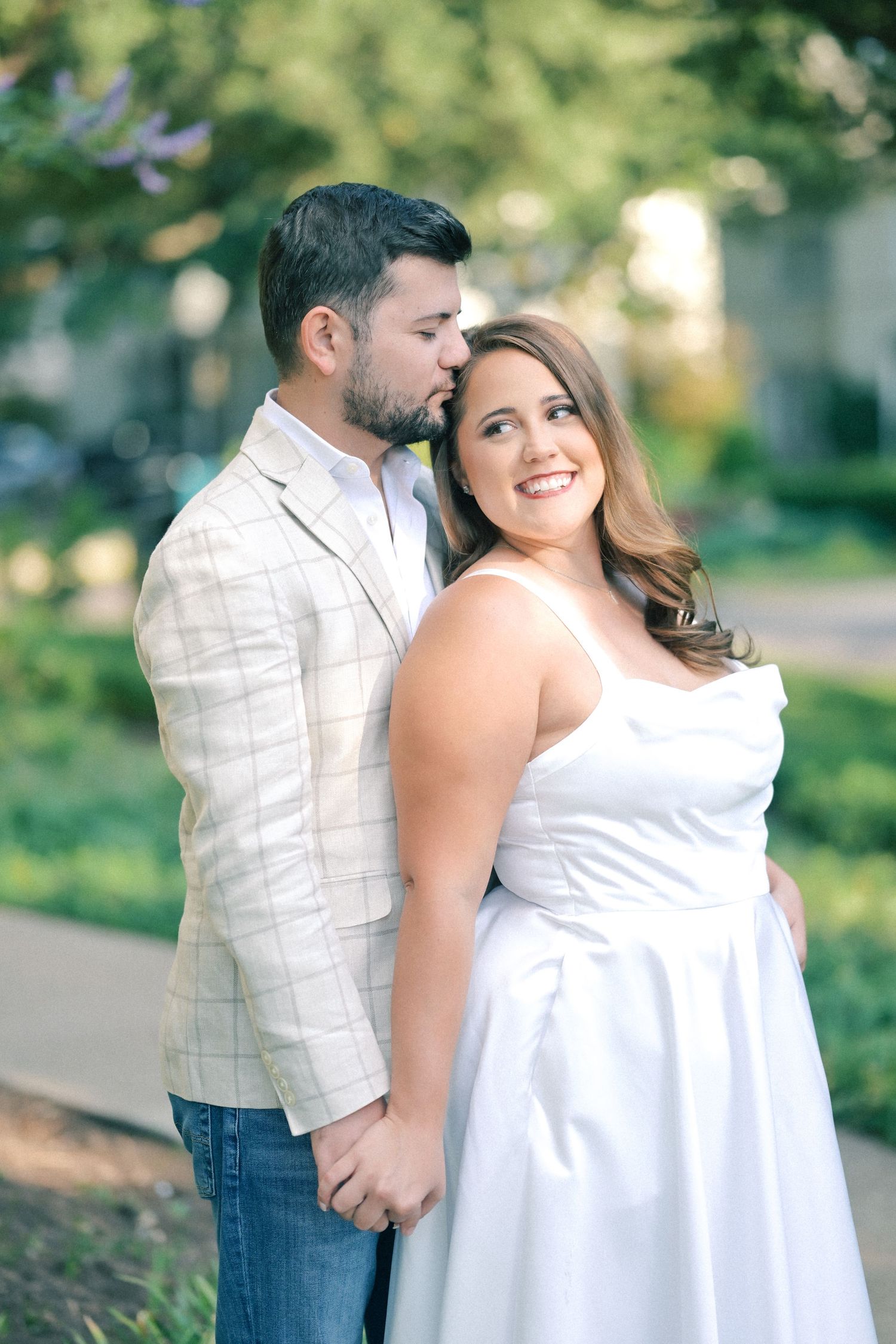 Smiling woman in a white dress holds hands with a man in a plaid blazer as he kisses her cheek in a sunny garden.