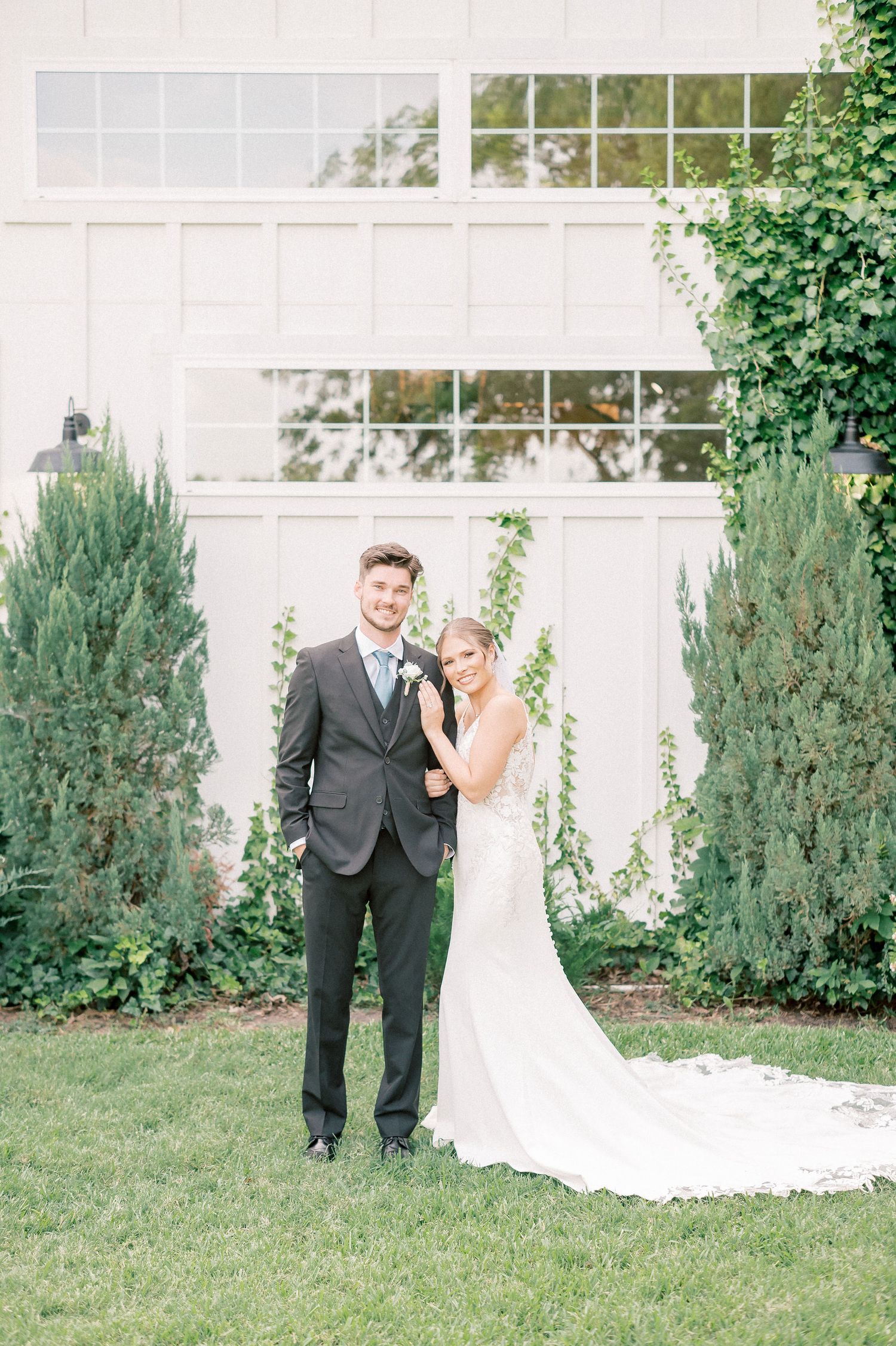 Full length wedding portraits taken outdoors against a white building with evergreen shrubs.