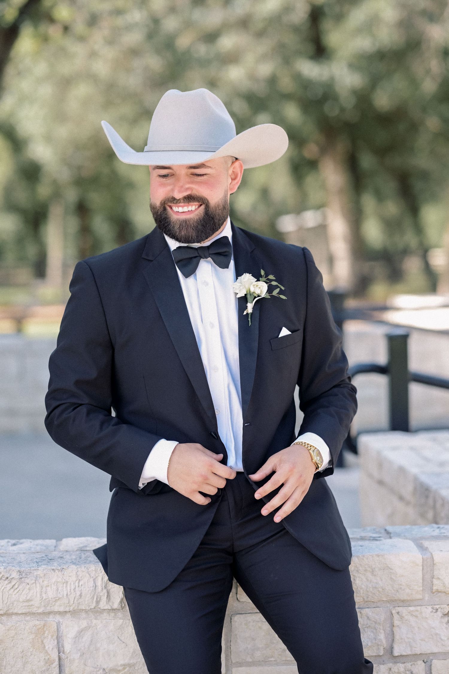 Smiling groom in black tuxedo with bow tie and white cowboy hat adjusting jacket outdoors.