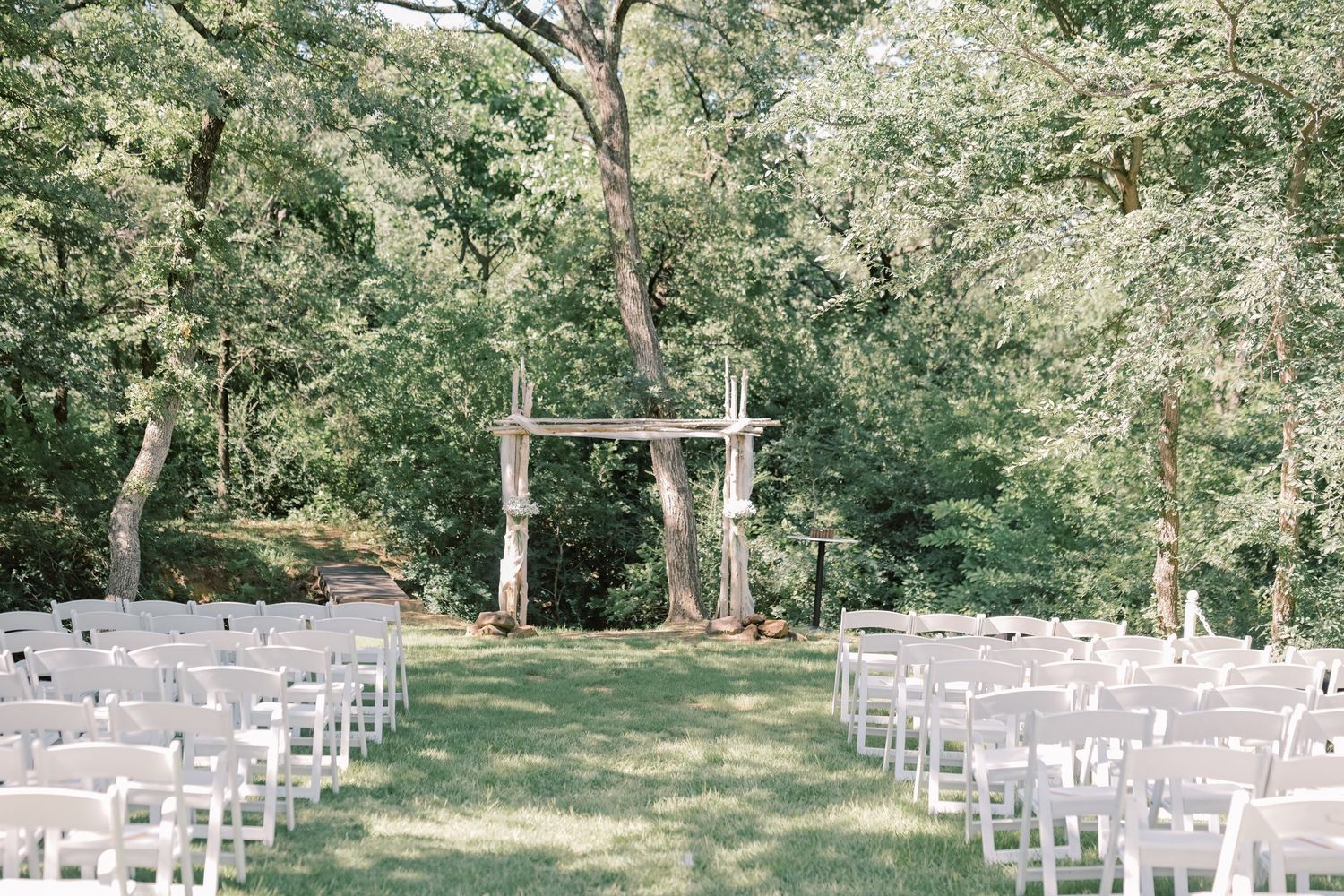 Outdoor wedding ceremony setup with white chairs and rustic wooden arch surrounded by tall green trees.