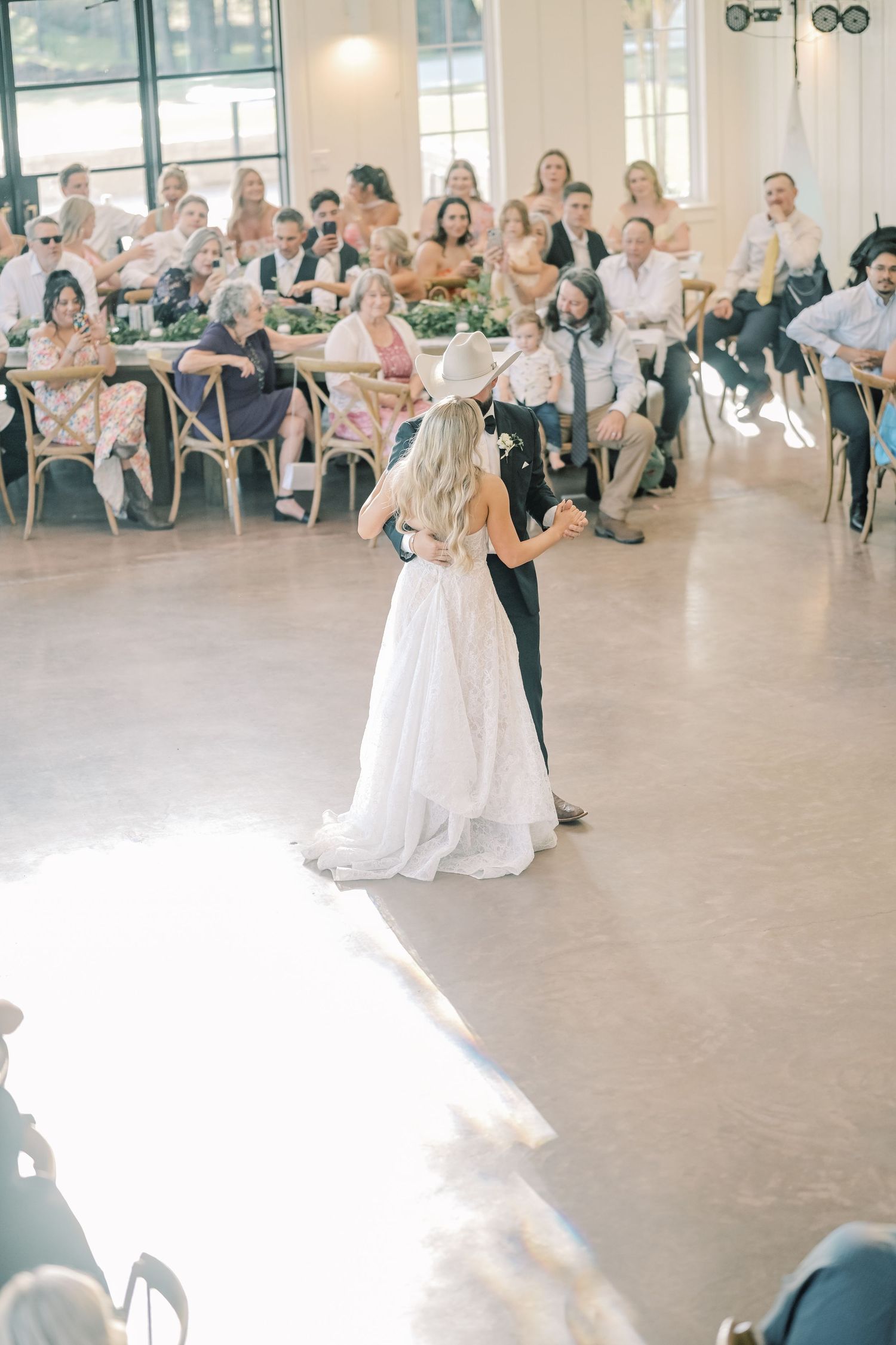 Bride and groom share first dance in bright reception hall surrounded by seated wedding guests.