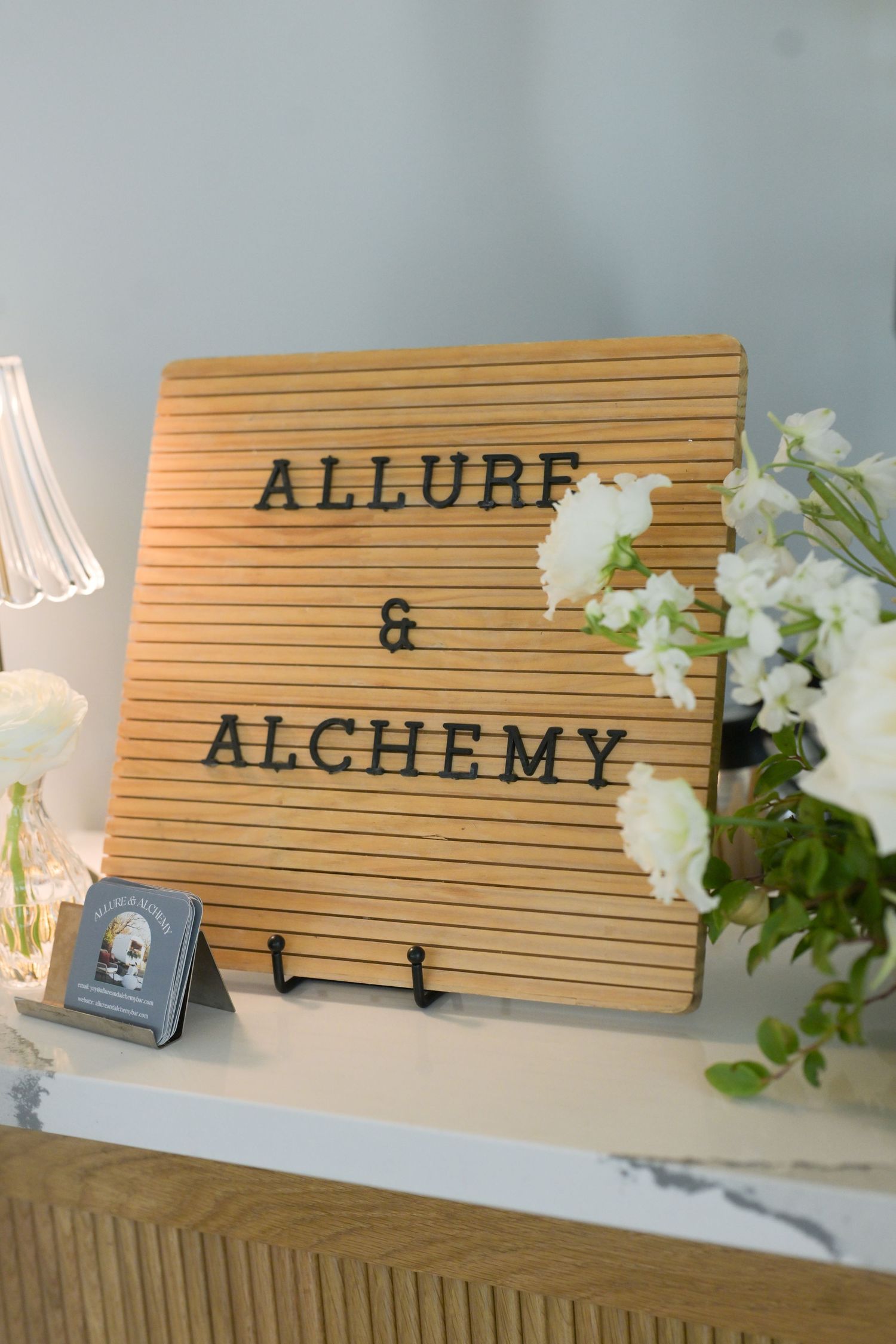 Wooden letter board sign reading Allure and Alchemy displayed with white flowers at a wedding venue.