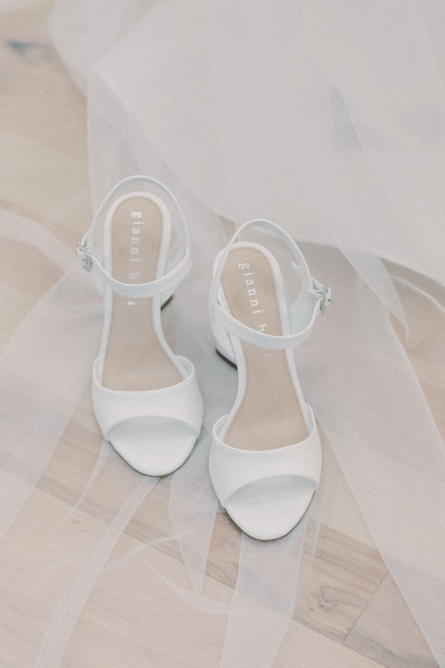 White block heel bridal sandals with ankle strap resting on sheer wedding veil on light wood floor.