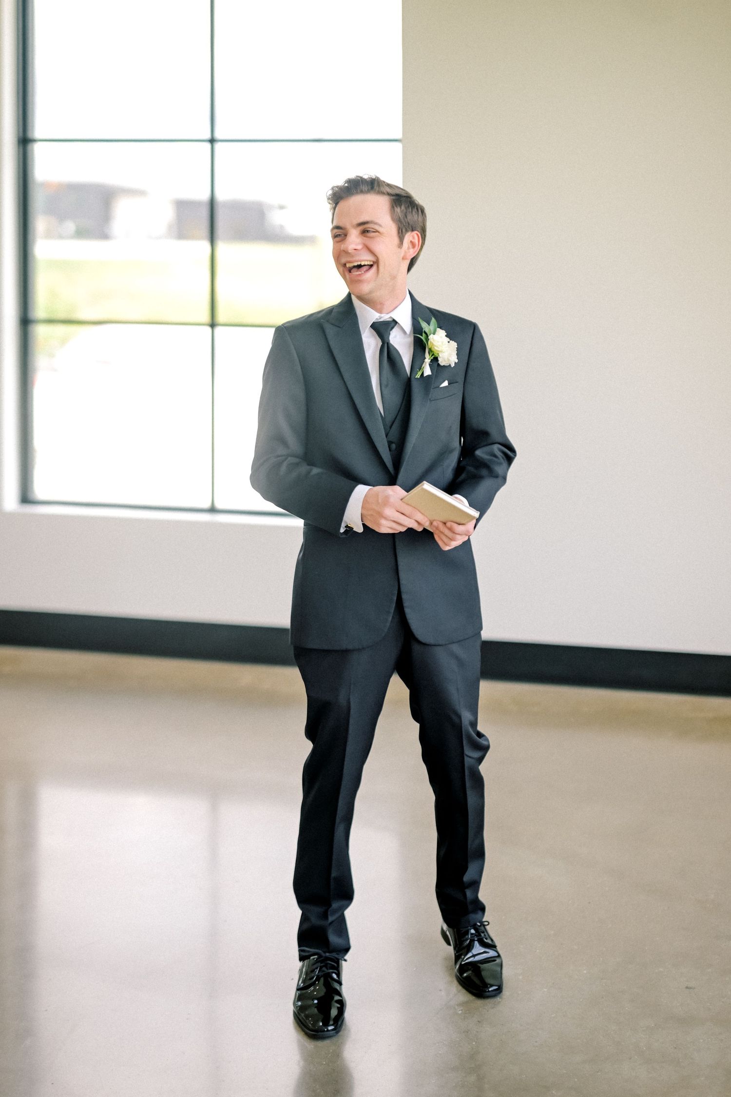 Smiling groom in a dark suit with boutonniere holding notes beside a large window in a modern venue.