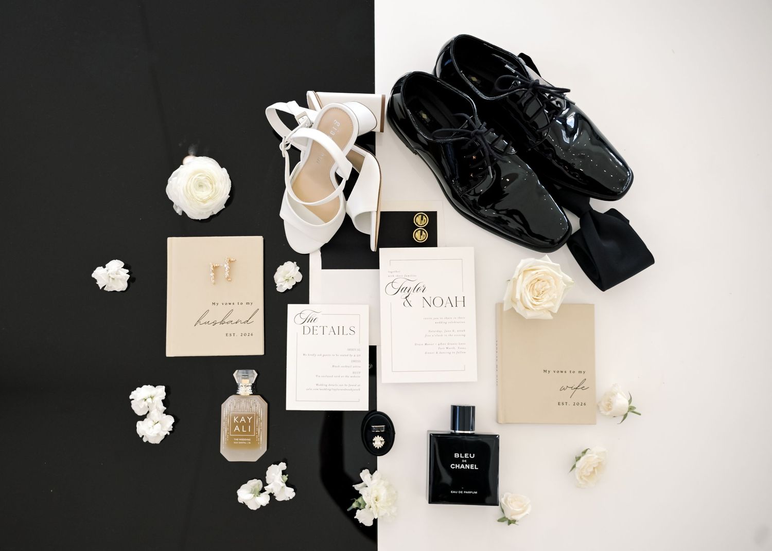 Elegant wedding flat lay with black patent shoes, white heels, perfume, invitations, and white roses on black and white background.