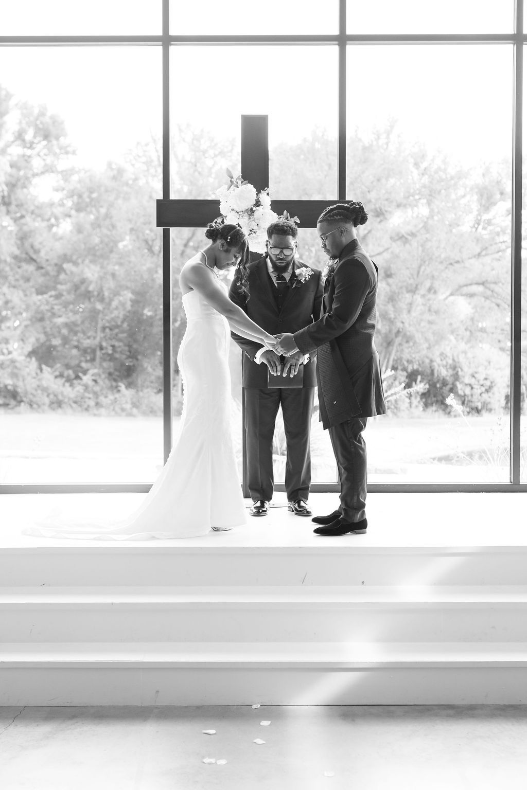 Black and white wedding photo of bride and groom holding hands at altar with cross and officiant.
