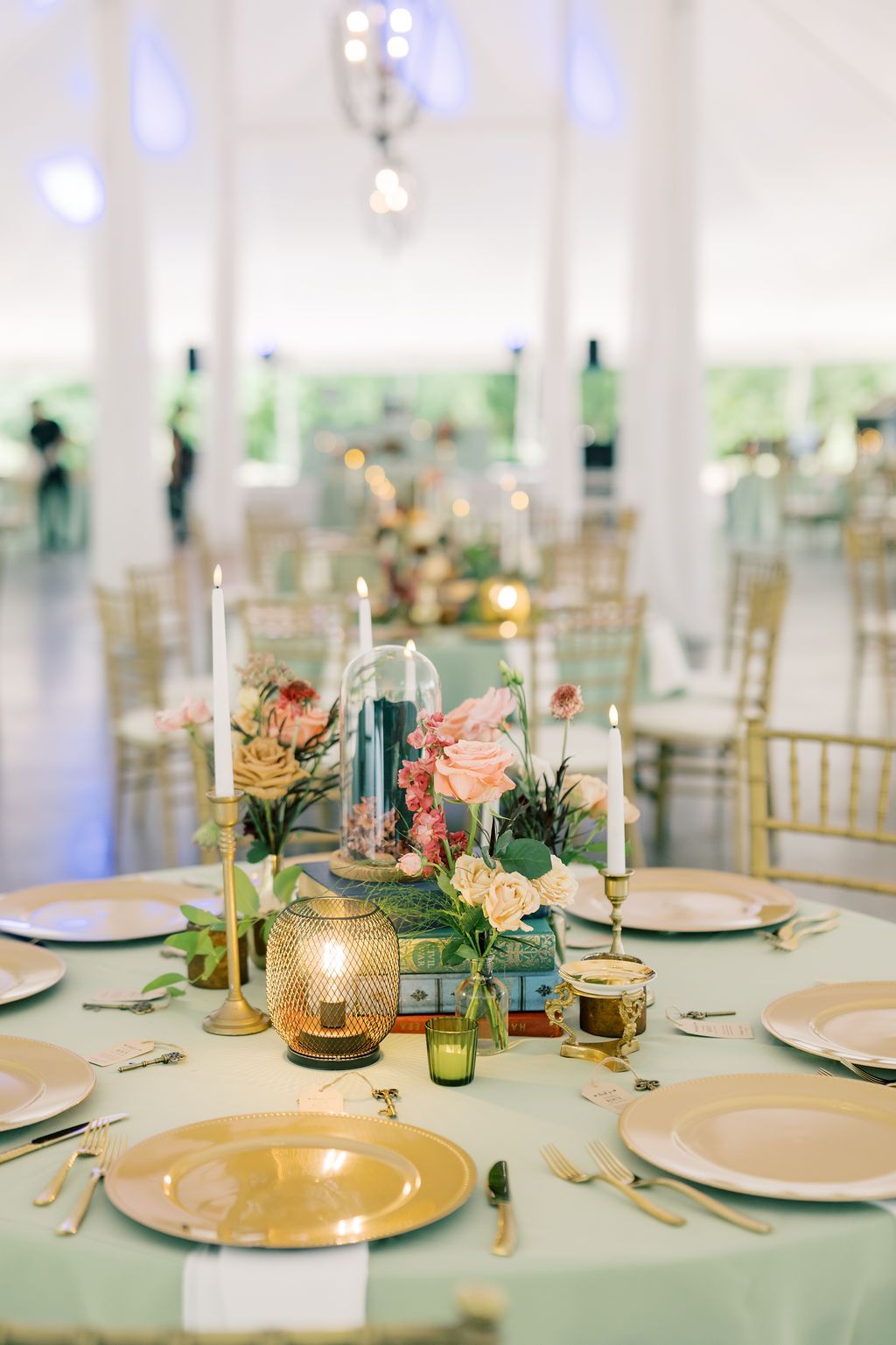 Elegant wedding reception table with mint green linen, gold chargers, floral centerpiece, candles, and chiavari chairs in a white tent.