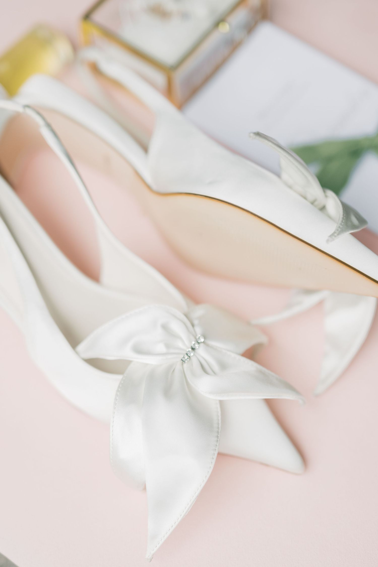 Elegant white satin bridal flats with a large bow detail on a soft pink background with wedding accessories.