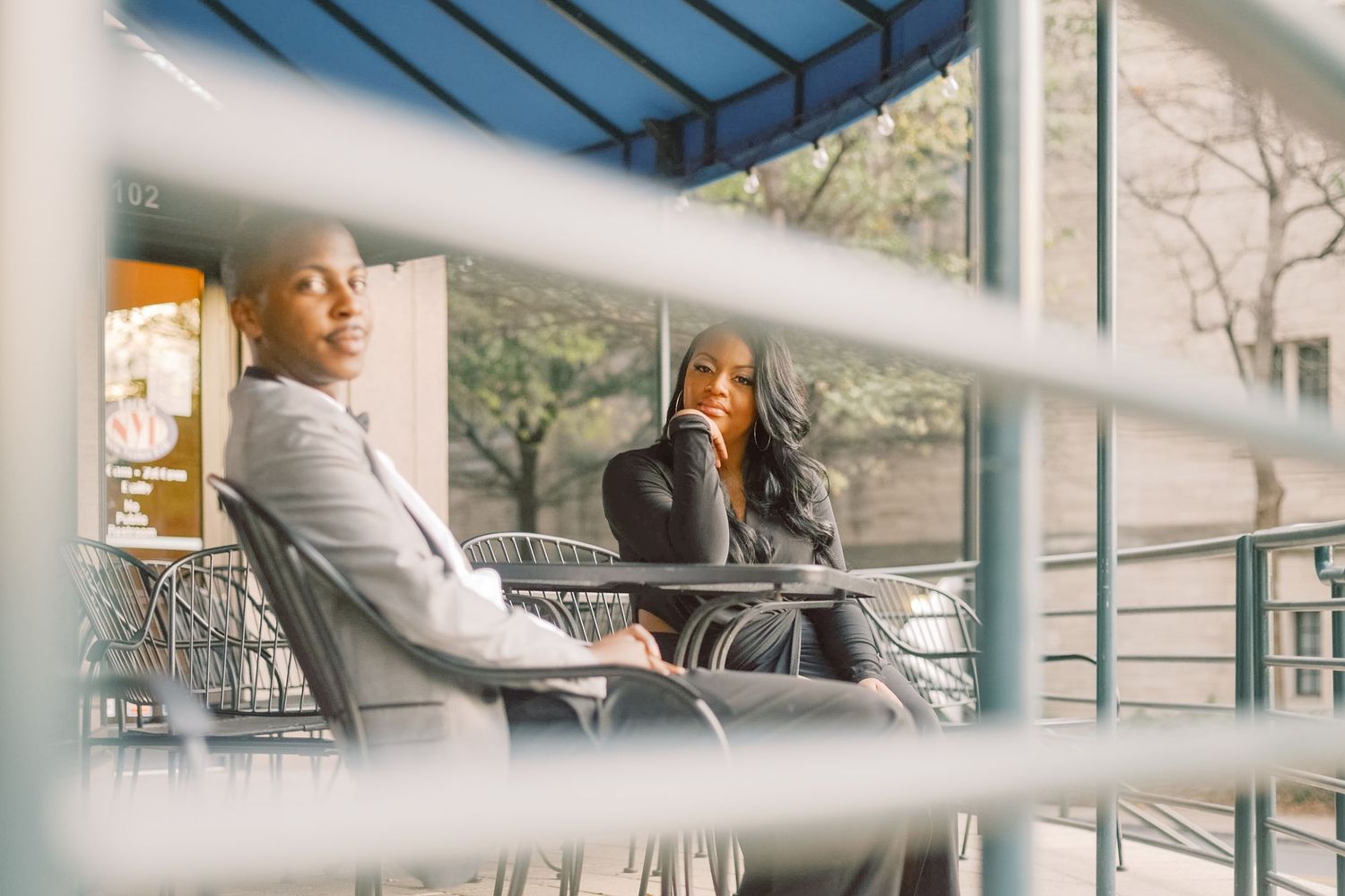 A man and woman sit together at an outdoor cafe table, engaged in conversation behind metal railings.
