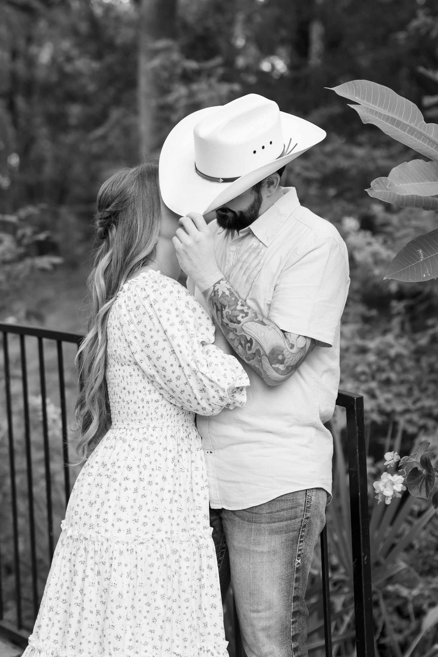 Black and white photo of couple kissing, man wearing cowboy hat with tattoos, near garden fence.