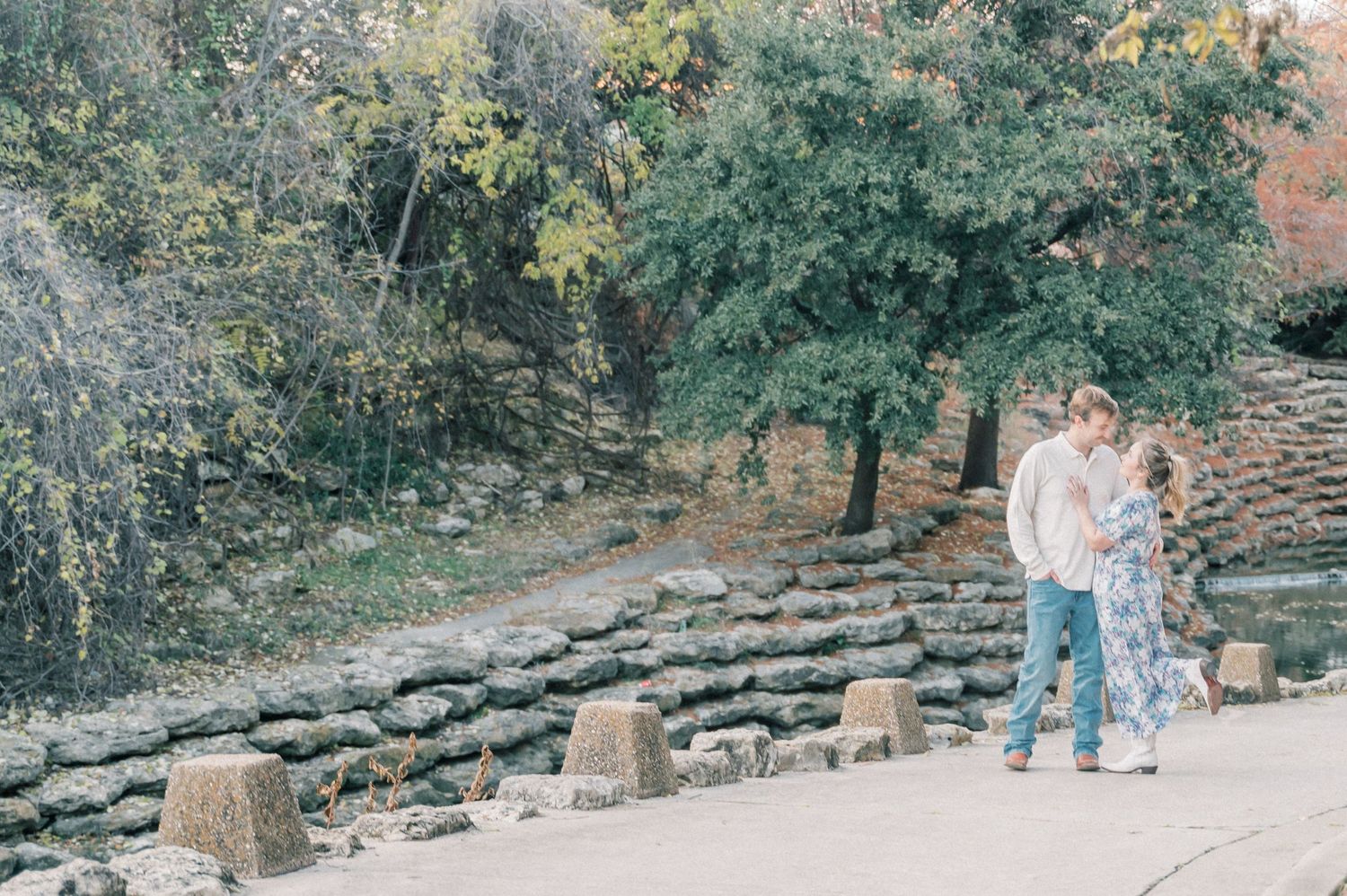 Couple walking along stone wall path in autumn garden with fallen leaves and rustic outdoor scenery.