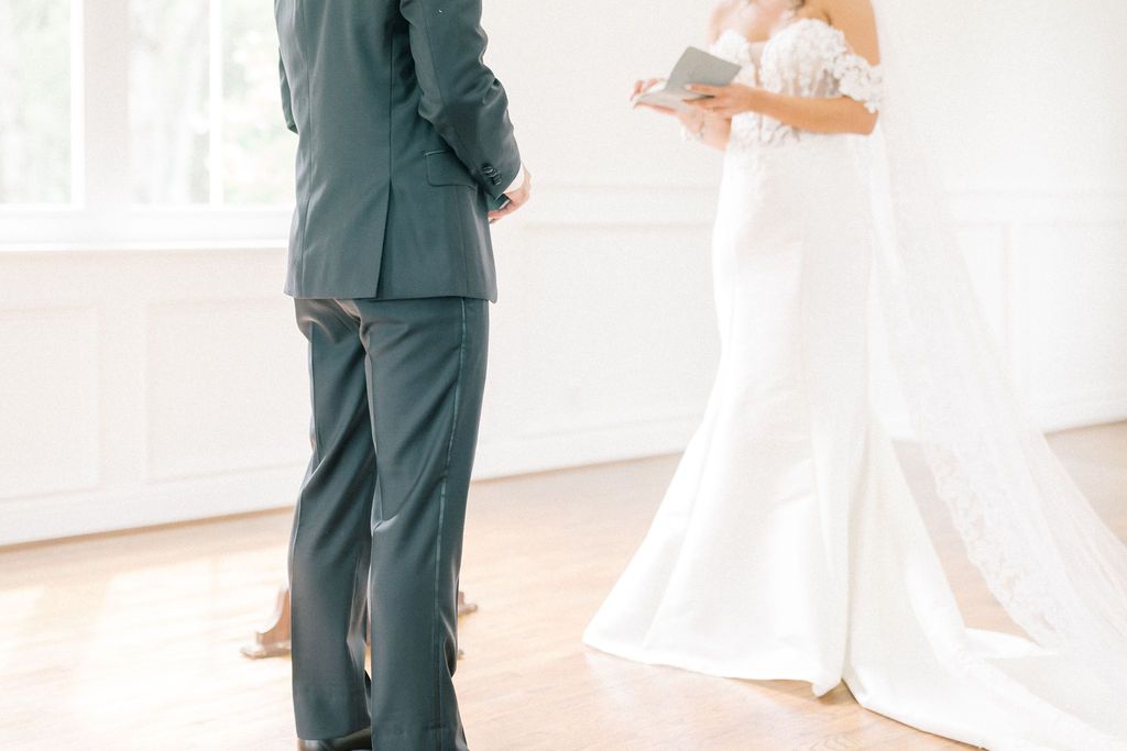 A bride in a white gown reads wedding vows to a groom in a gray suit during an intimate ceremony.