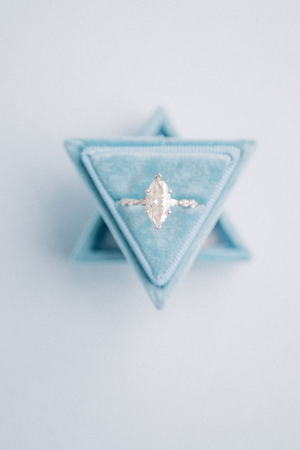 Marquise diamond engagement ring in a light blue triangular velvet ring box on soft background.