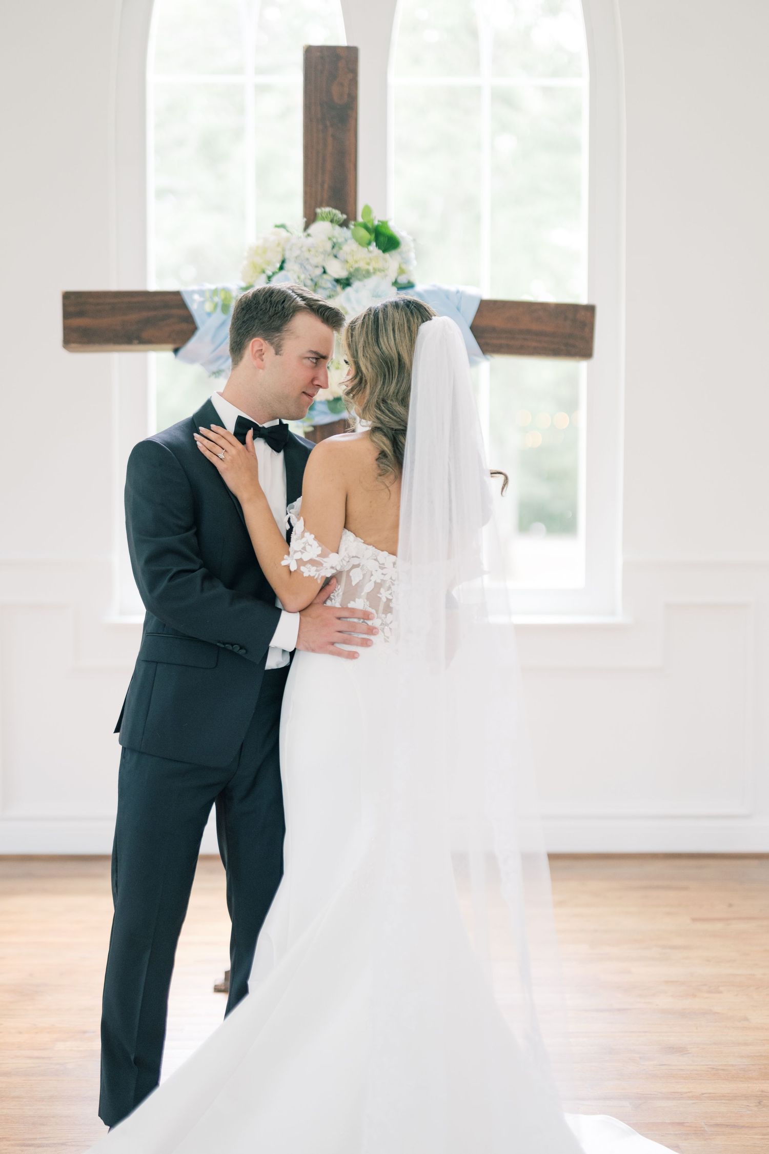 Bride and groom embrace before wooden cross altar with floral arrangement in bright white chapel.