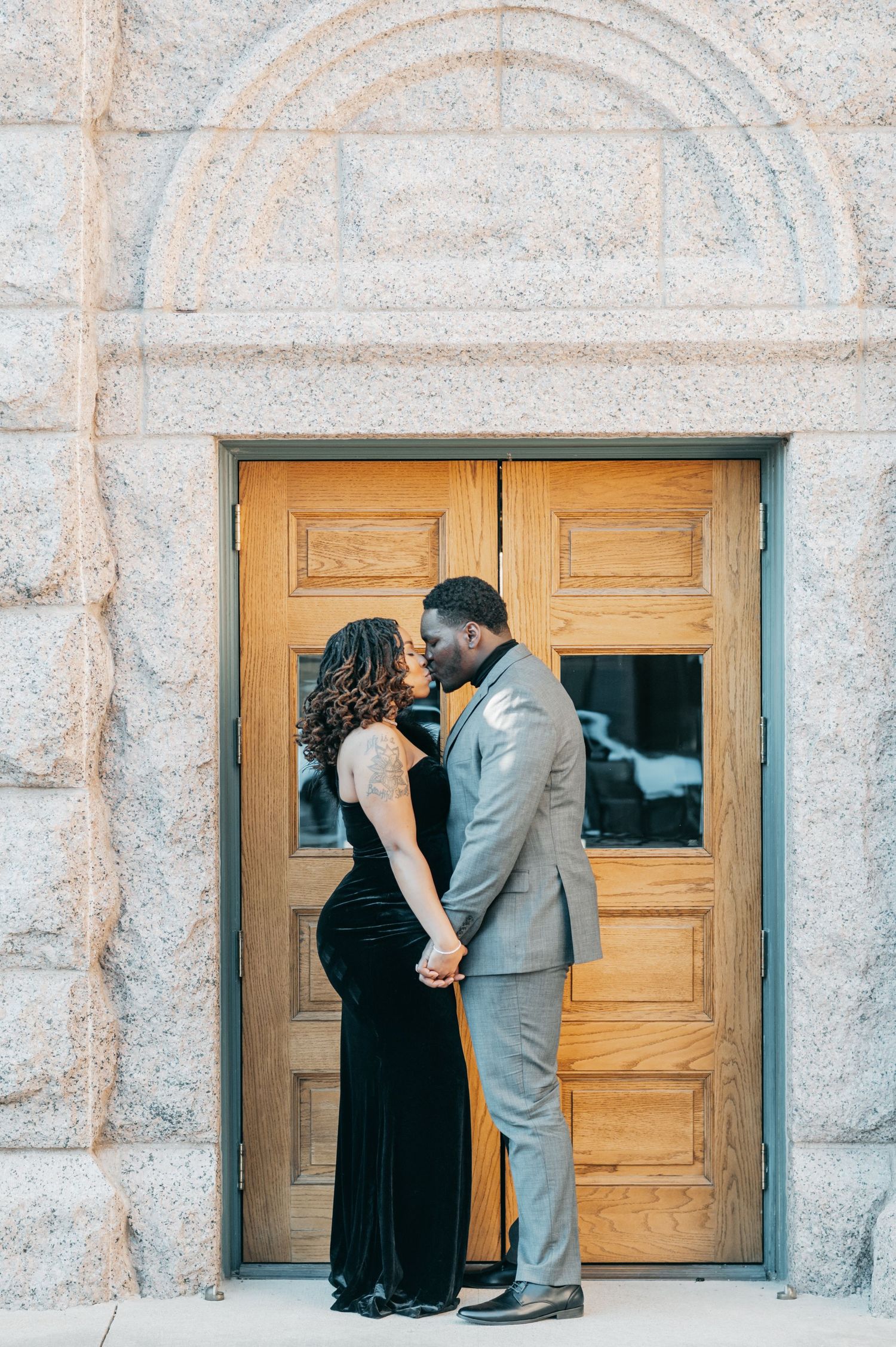 Couple sharing a romantic kiss in front of elegant wooden doors of a stone building, dressed in formal attire.
