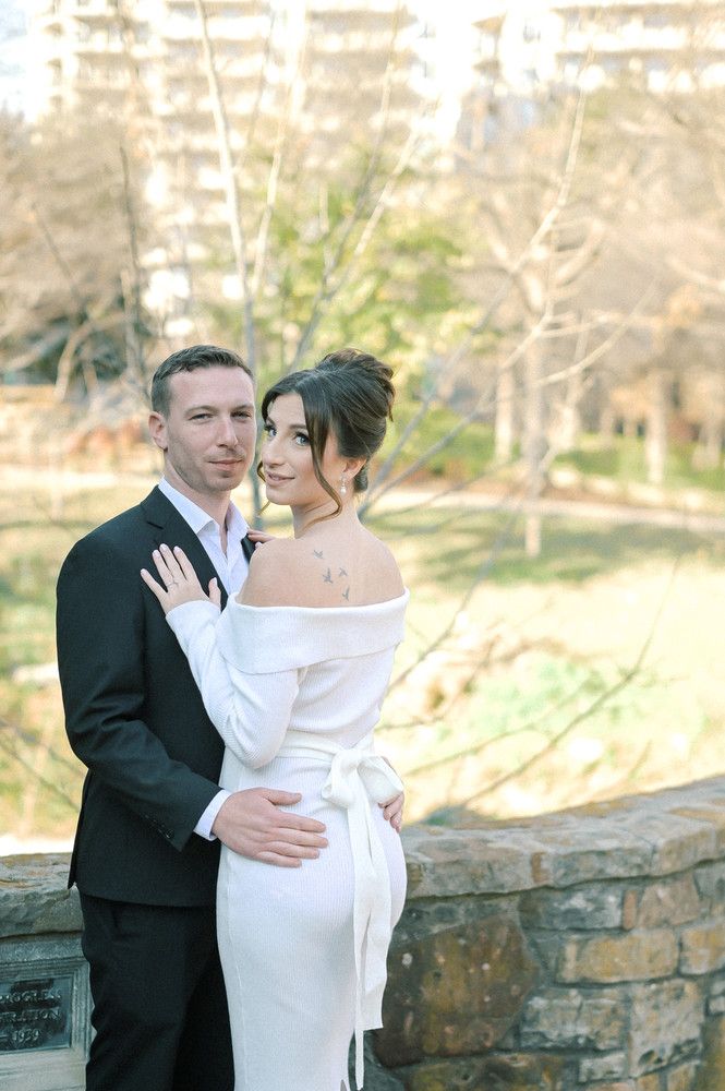 Bride in elegant off-shoulder white gown embraces groom in black suit near a stone wall in a park setting.
