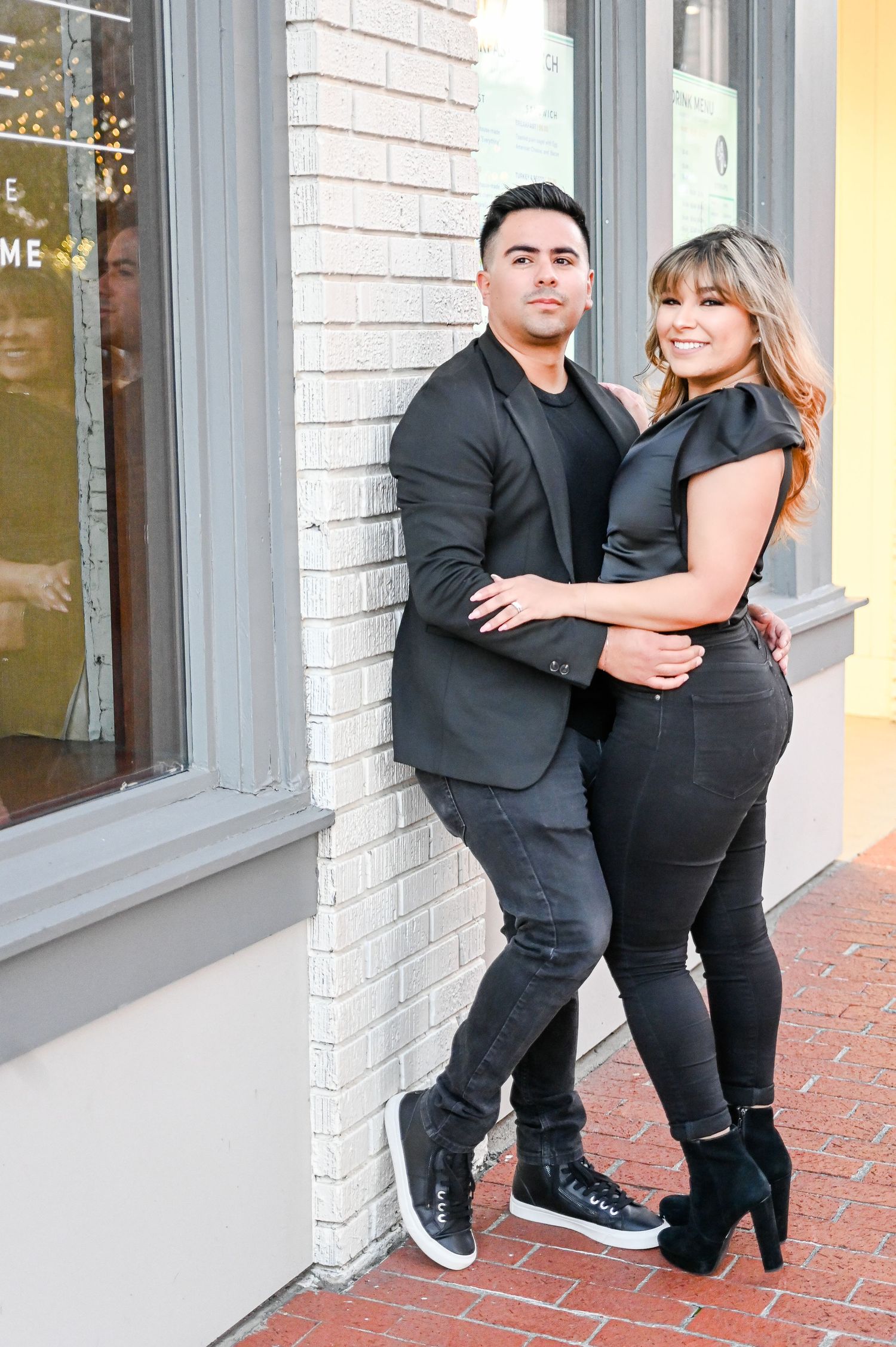 Stylish couple in all-black outfits embracing against a white brick wall.