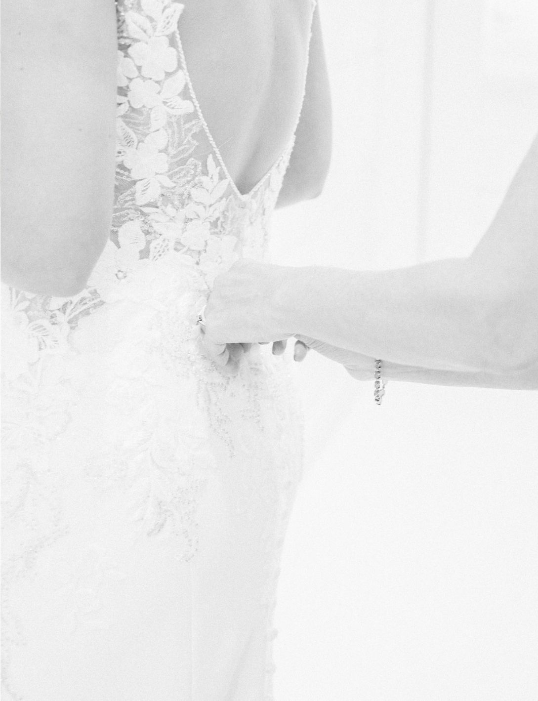 Black and white photo of a bride in a lace wedding dress being buttoned up from behind.
