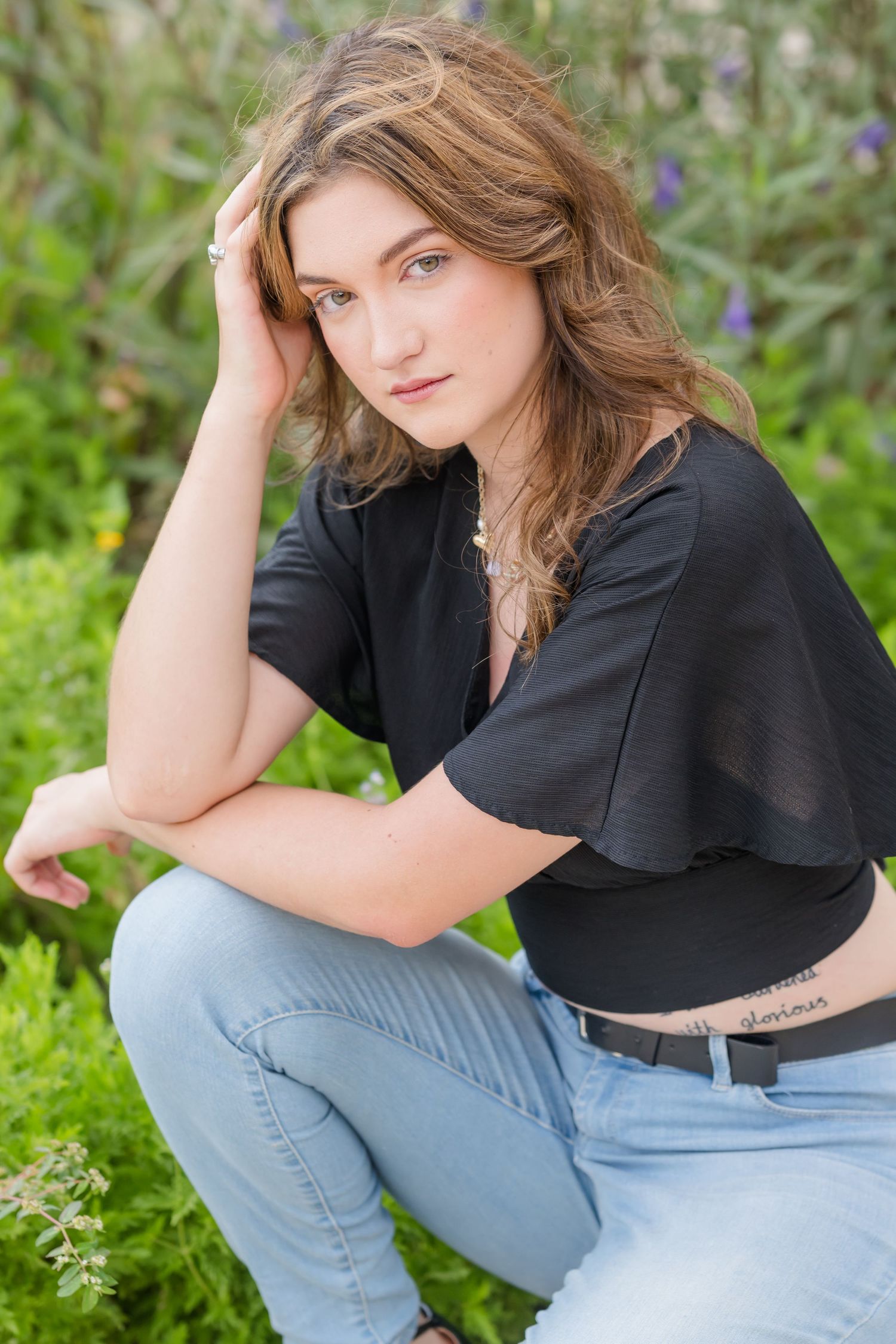 Young woman with wavy brown hair in black top and jeans crouching outdoors among green plants.
